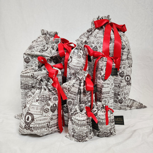 Set of ten reusable Christmas gift bags in Santas Mail. Bags are white with black Christmas postage stamps on them, tied with a red satin ribbon.