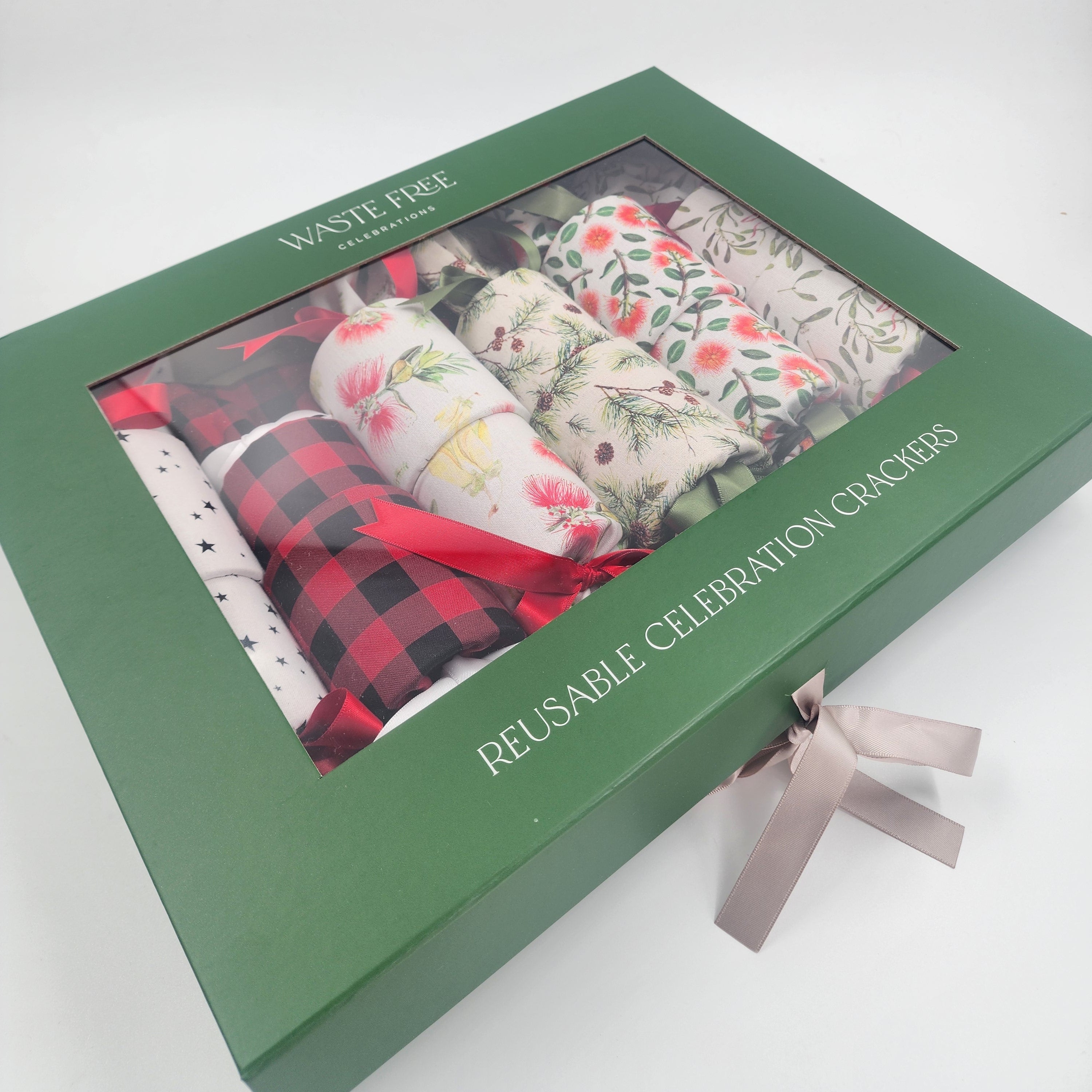 A green gift box labeled 'WASTE FREE' and 'REUSABLE CELEBRATION CRACKERS' with a clear window showing several rolled cotton cracker packs in different holiday prints, tied with a pale grey ribbon on a white background.