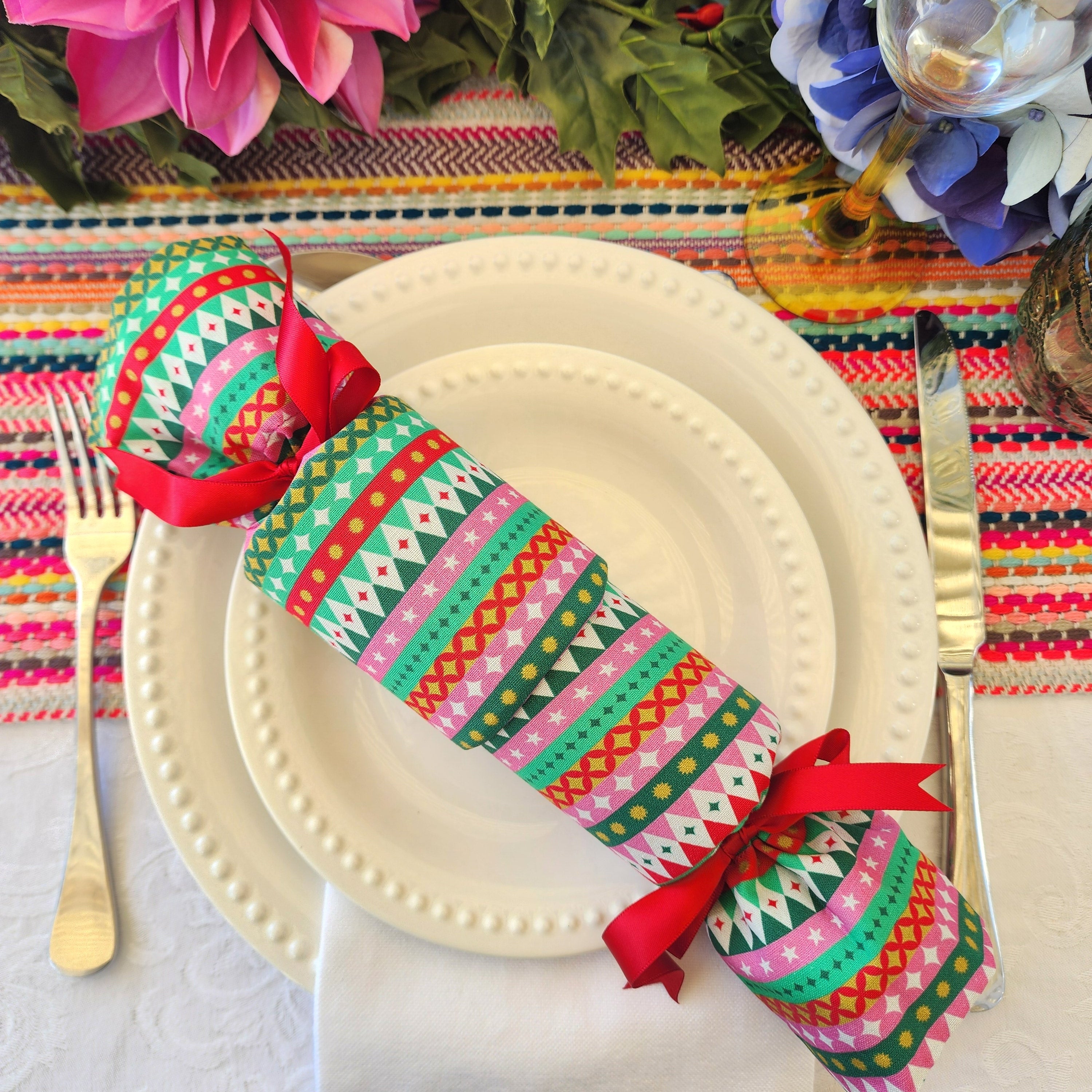 A festive Christmas table setting featuring a colorful patterned reusable cracker with red ribbons, placed on a white plate with gold-toned cutlery on a striped table runner.