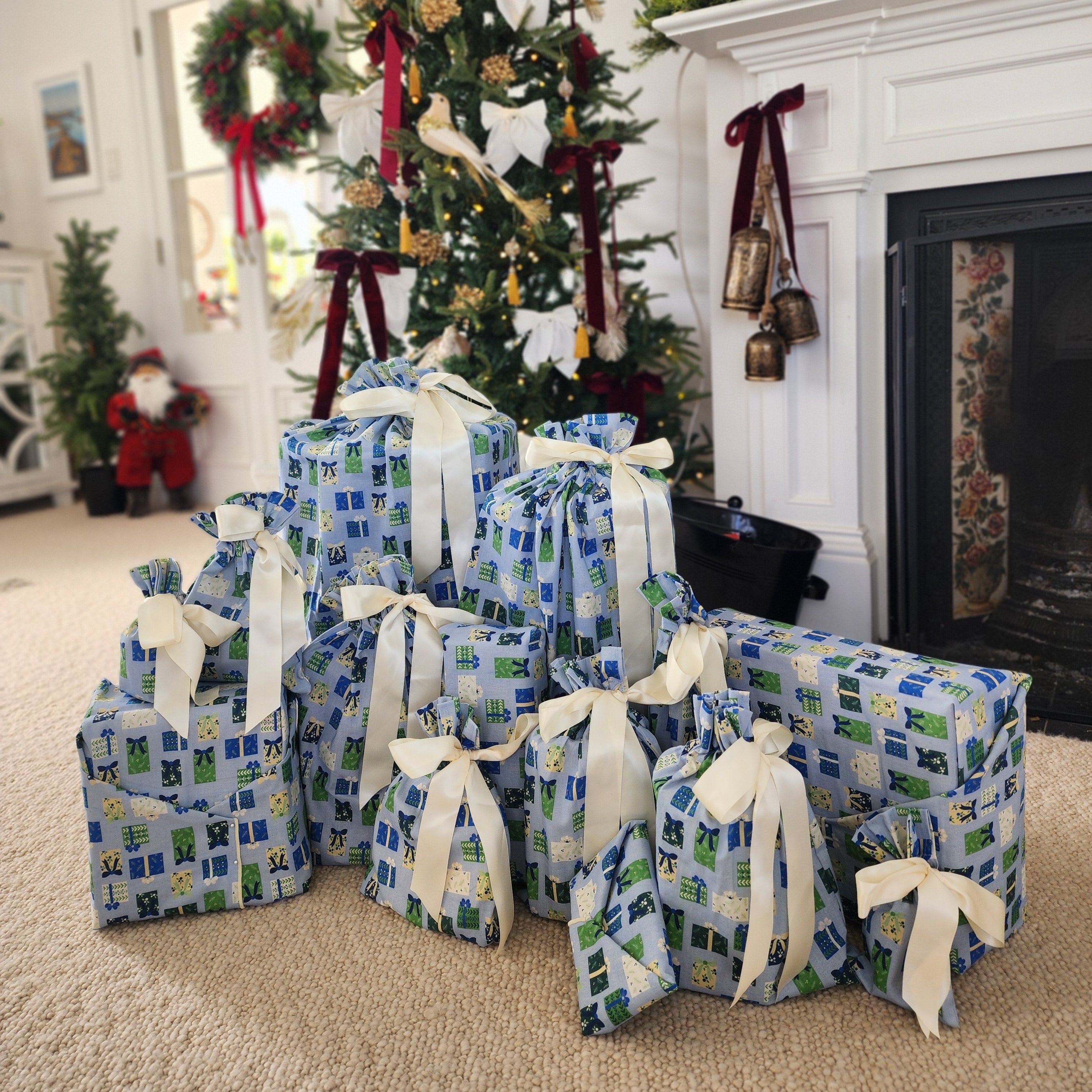 A group of wrapped Christmas gifts in blue and green patterned fabric with cream ribbons, arranged on a carpeted floor in front of a decorated Christmas tree and fireplace.