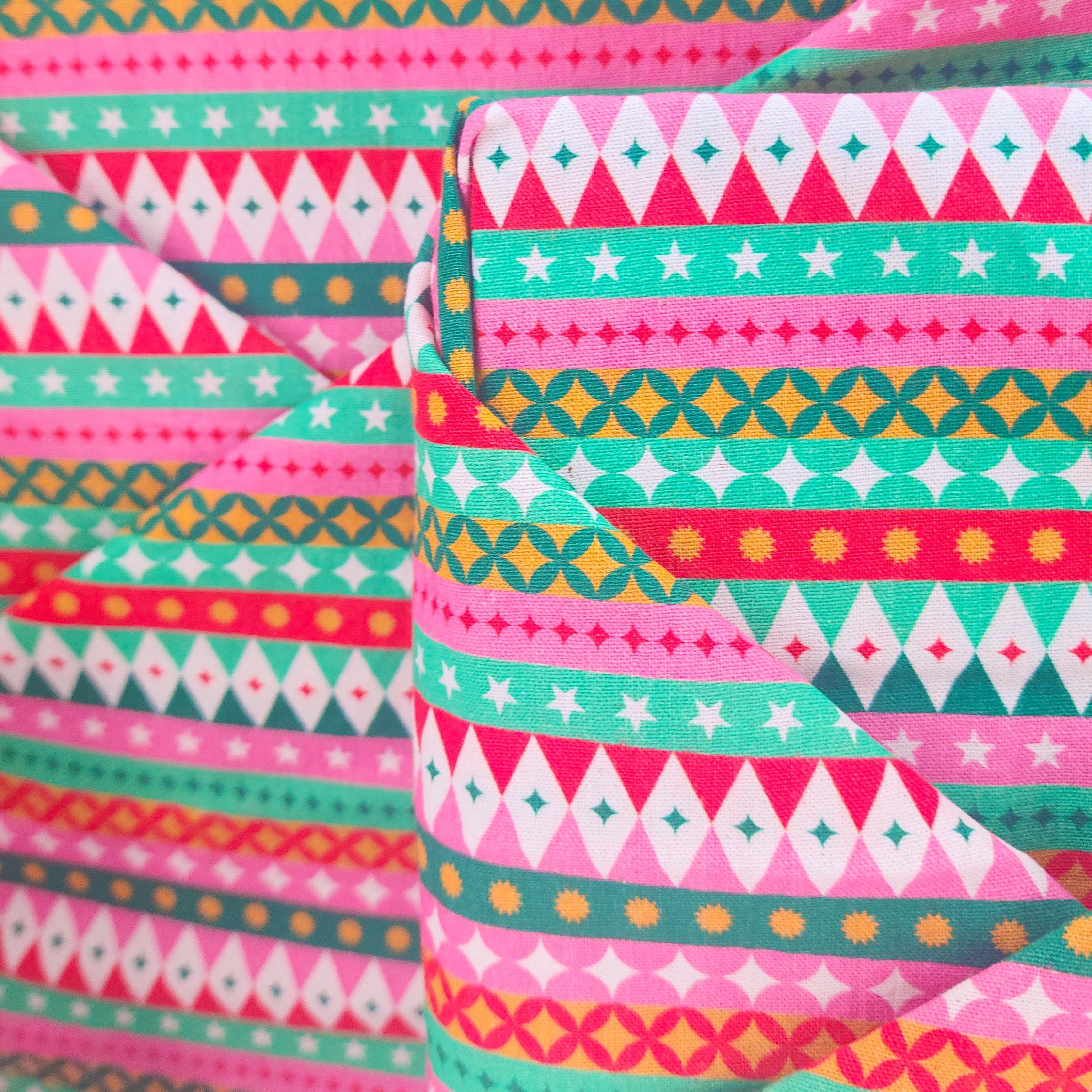 A close-up of folded and stacked cotton-wrapped gifts featuring a colorful geometric pattern with pink, teal, white, orange, and yellow stripes, triangles, stars, and dots.