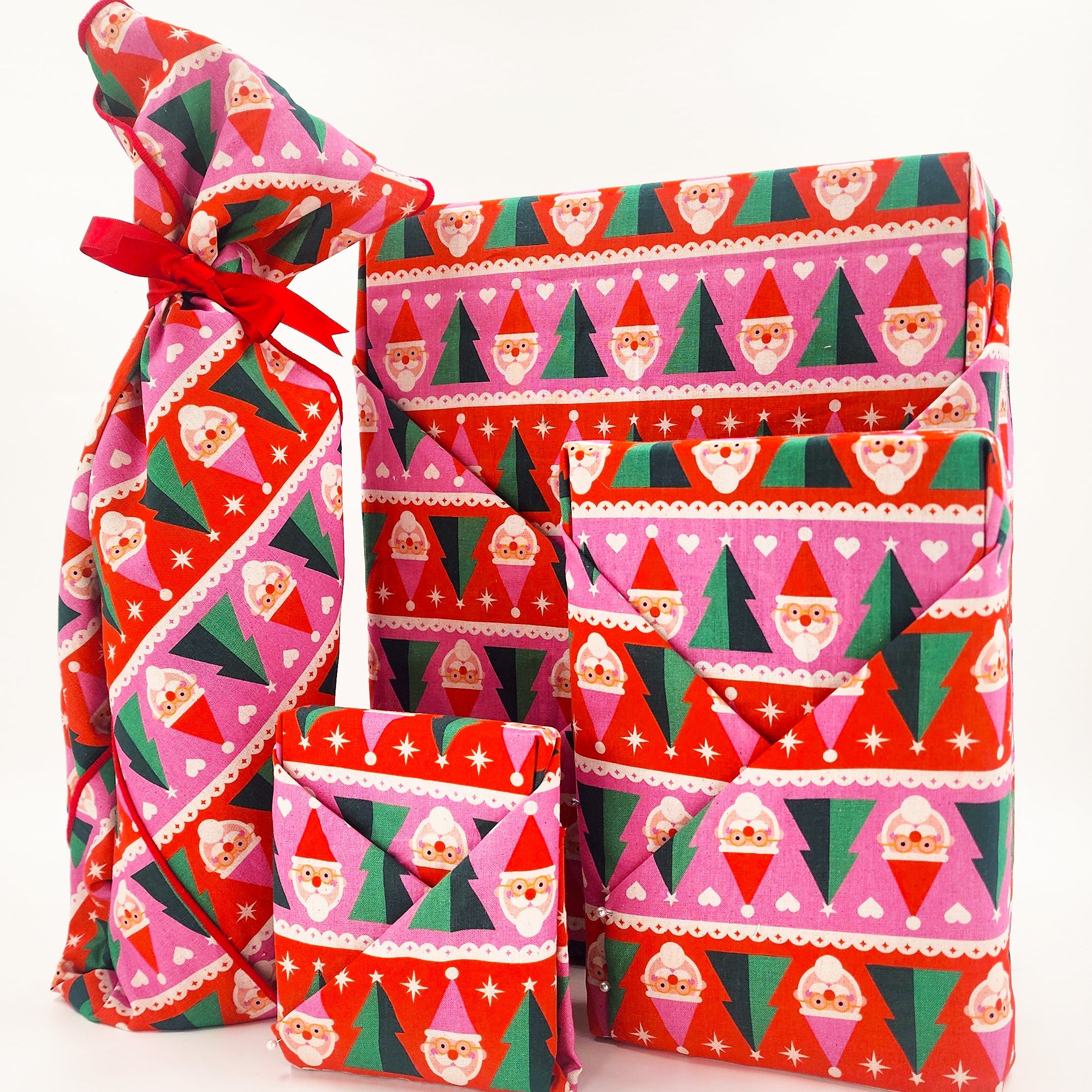 Three furoshiki wrapped gift boxes in different sizes with a festive Christmas pattern featuring Santa Claus faces, Christmas trees, and hearts in red, pink, green, and white, set against a plain white background.