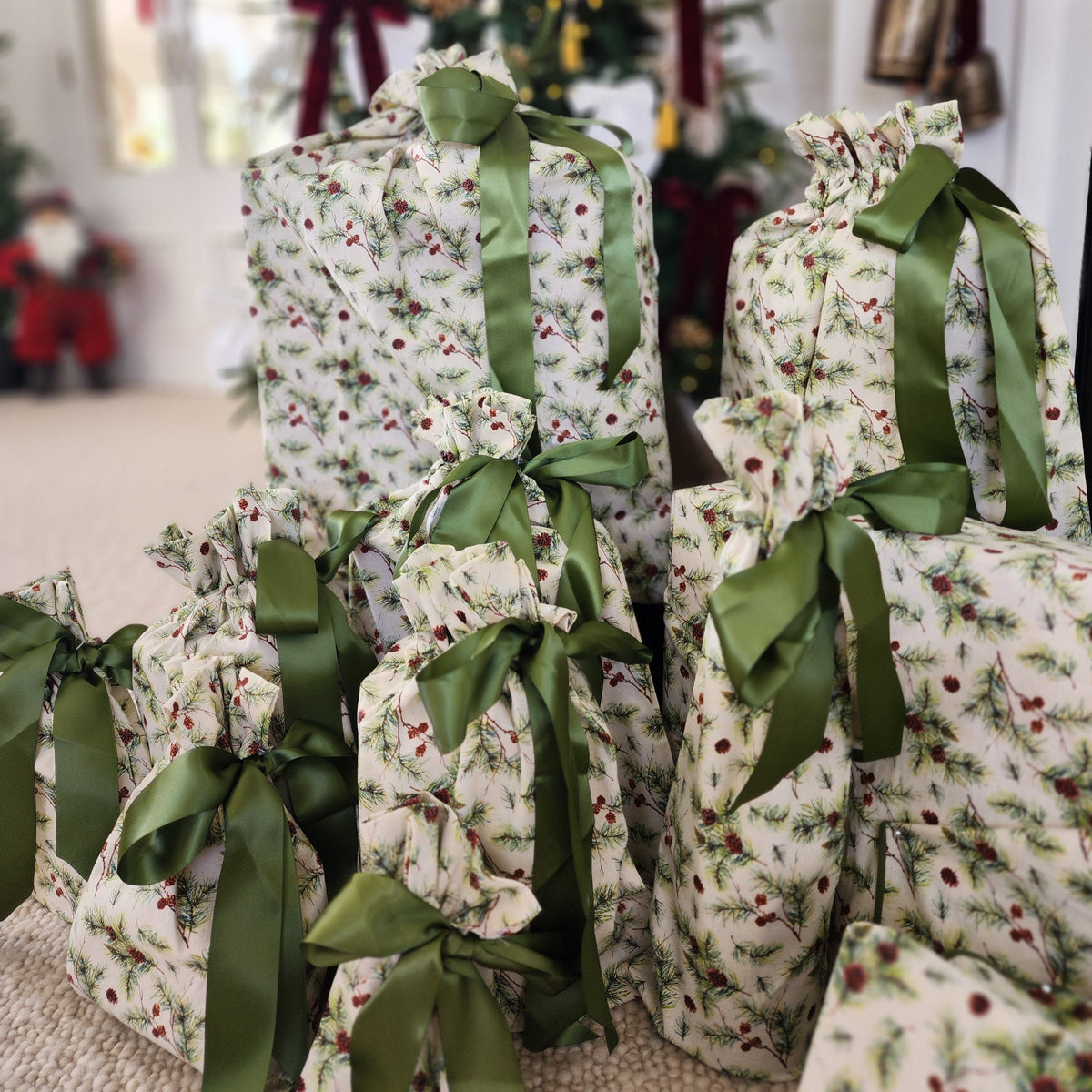 A group of Christmas wrapped gift bags in a holly and ivy pattern, tied with olive green satin ribbons, arranged on a carpeted floor in front of a decorated Christmas tree.