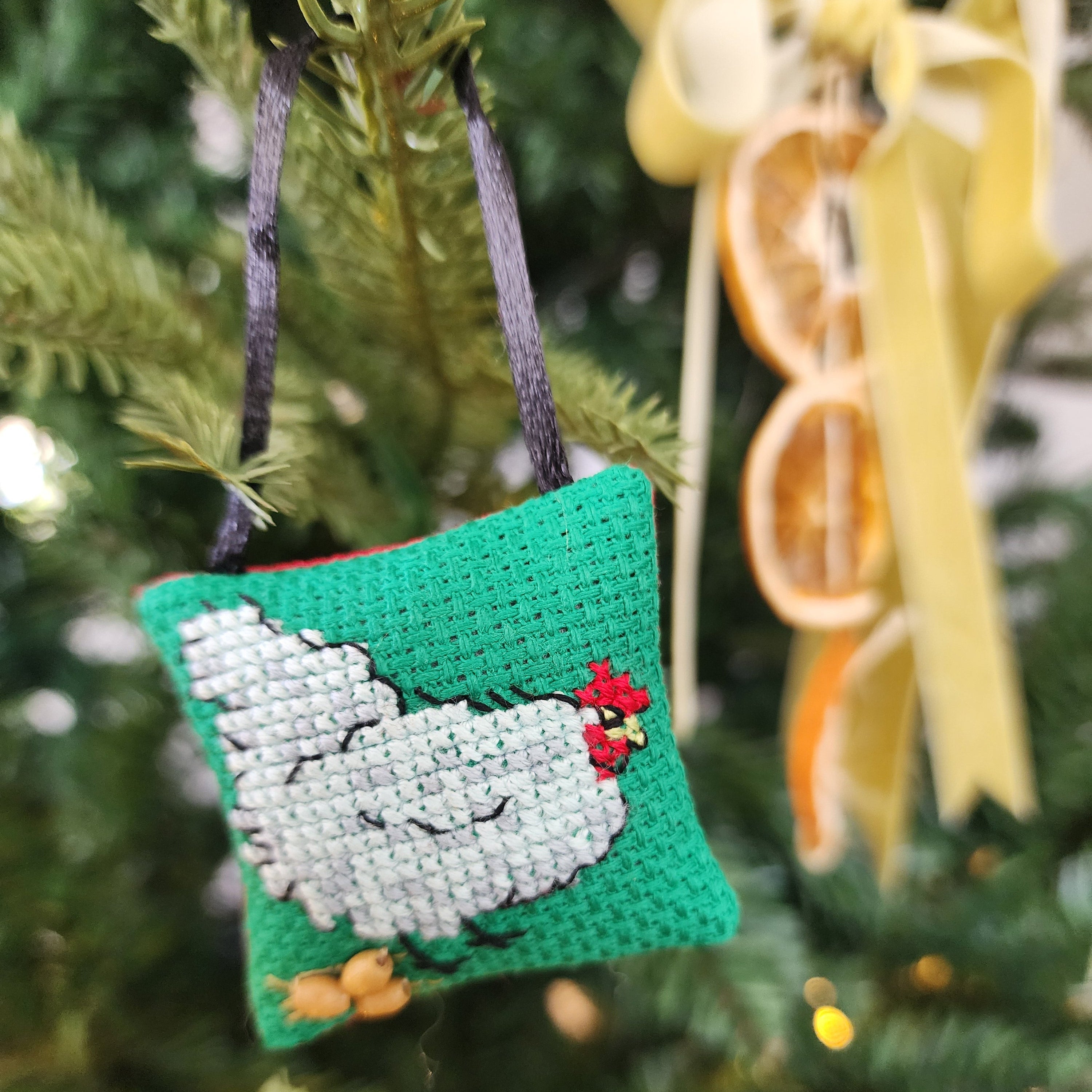 Cross-stitched ornament of a chicken laying eggs on green background, hanging on a Christmas tree.