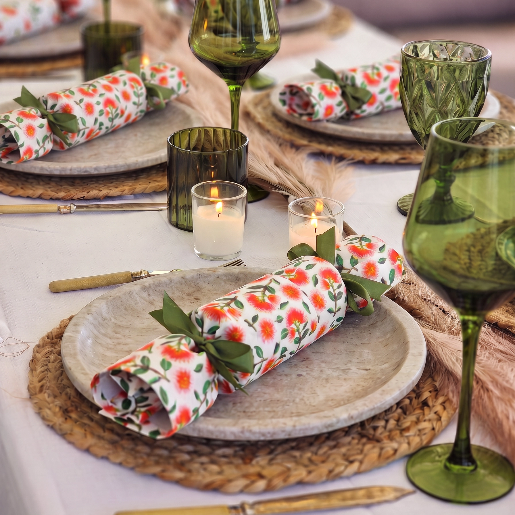 A styled table setting with green glass wine glasses, woven placemats, white tablecloths, lit candles, and reusable christmas crackers in a pohutukawa print tied with green bows.