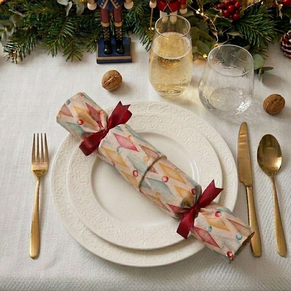 A festive Christmas table setting featuring a reusable Christmas cracker with a colorful geometric pattern and red ribbon, placed on a white plate. The setting includes gold-toned cutlery, glasses of champagne, nutcrackers, pine branches, and candles.