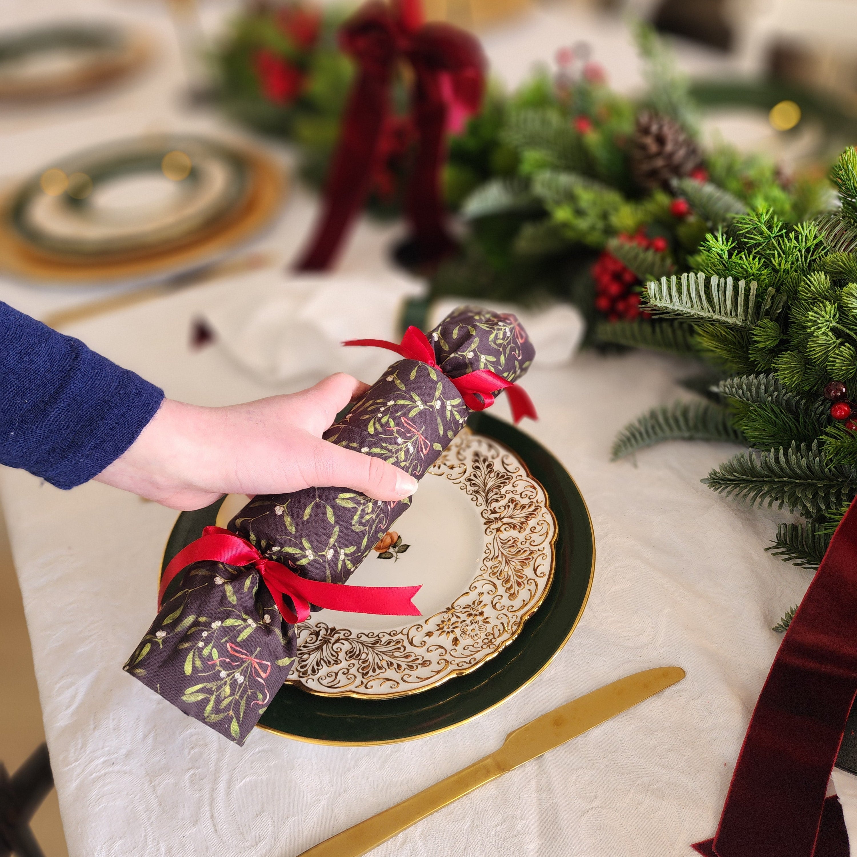 A hand wearing a navy blue sleeve holds a wrapped Christmas cracker ornament decorated with a gold and purple pattern and tied with a red ribbon, set on a decorated holiday table with gold cutlery, plates, and greenery.