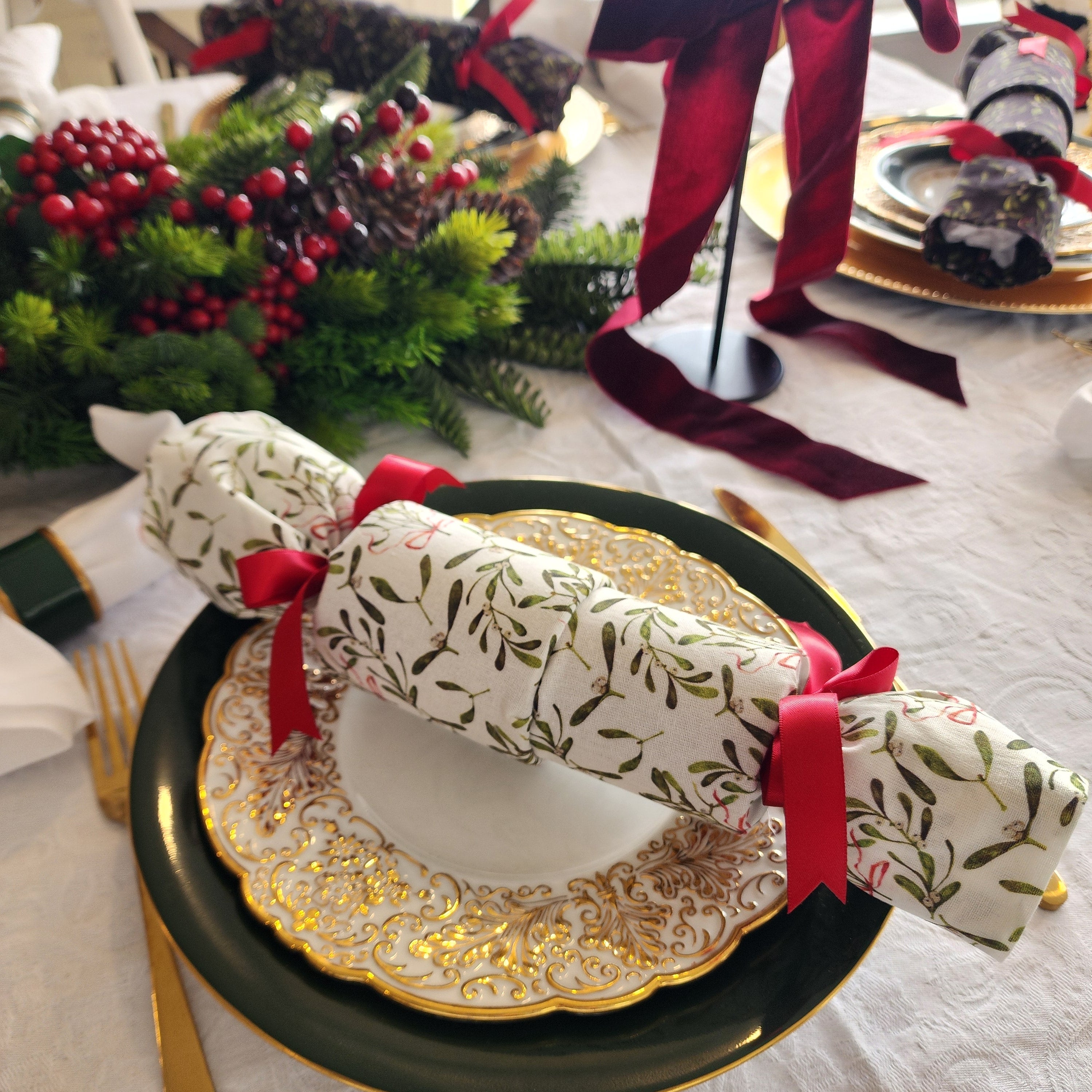 A styled holiday table setting featuring a white reusable cracker with a green leaf pattern and red ribbon bow, layered over gold and dark green plates on a white tablecloth.