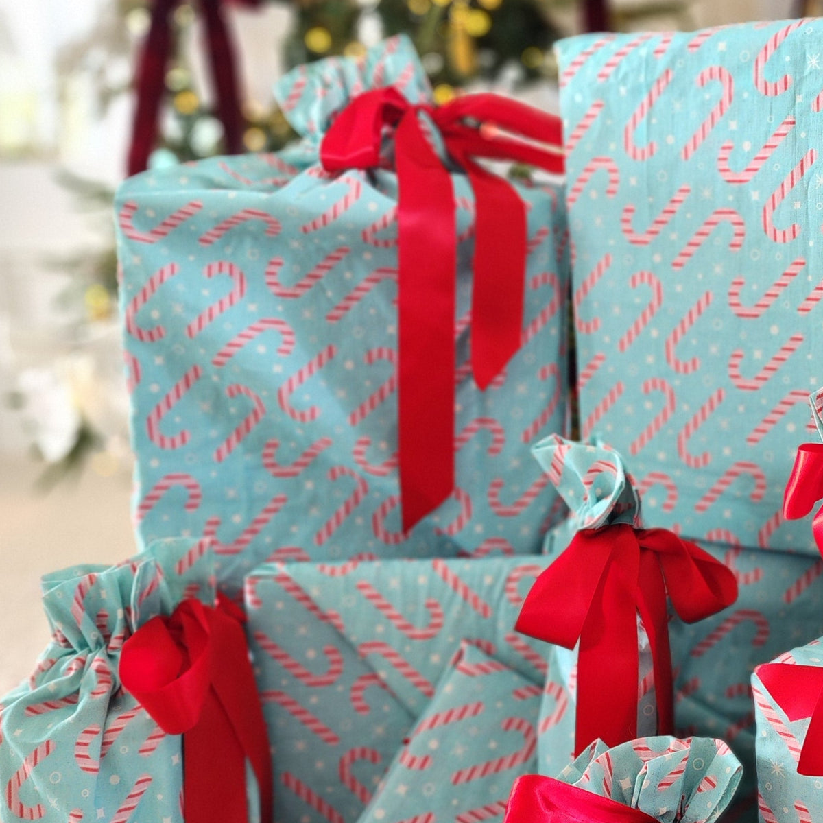 A stack of Christmas wrapped gifts in light blue cotton bags and furoshiki with a red candy cane pattern, tied with bright red ribbons, set on a carpeted floor in front of a decorated Christmas tree and fireplace.