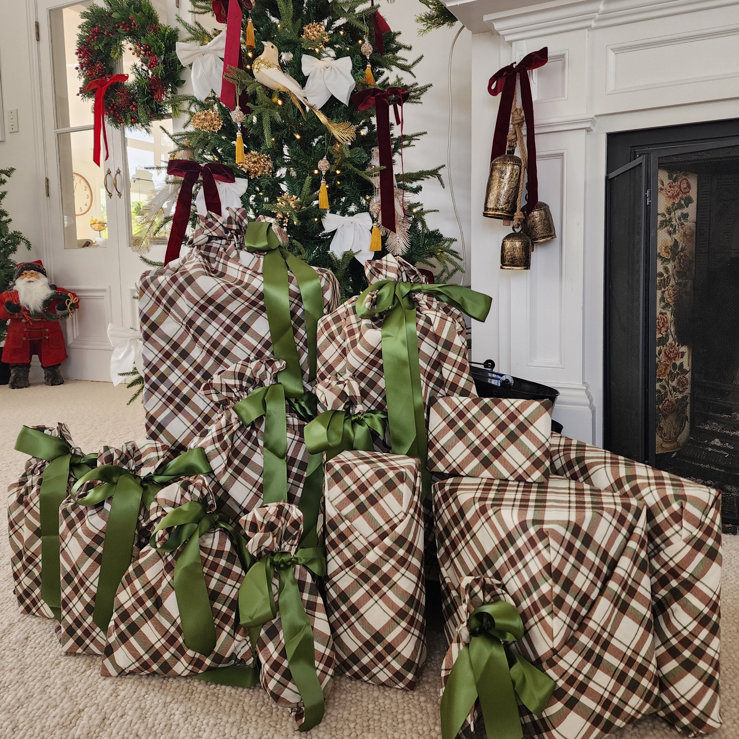 A group of Christmas wrapped gift boxes in brown and beige plaid cotton with green ribbons, gathered under a decorated Christmas tree in a living room next to a fireplace.
