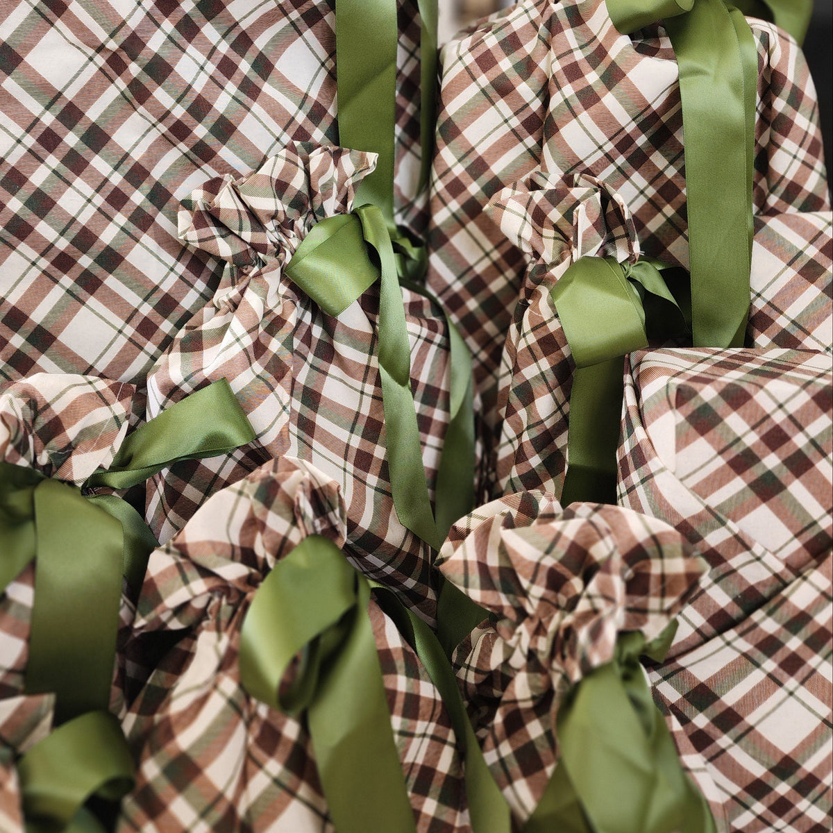 A group of Christmas wrapped gifts in brown, beige, and black plaid cotton with sage green satin ribbons tied in bows, set against a softly blurred holiday background.