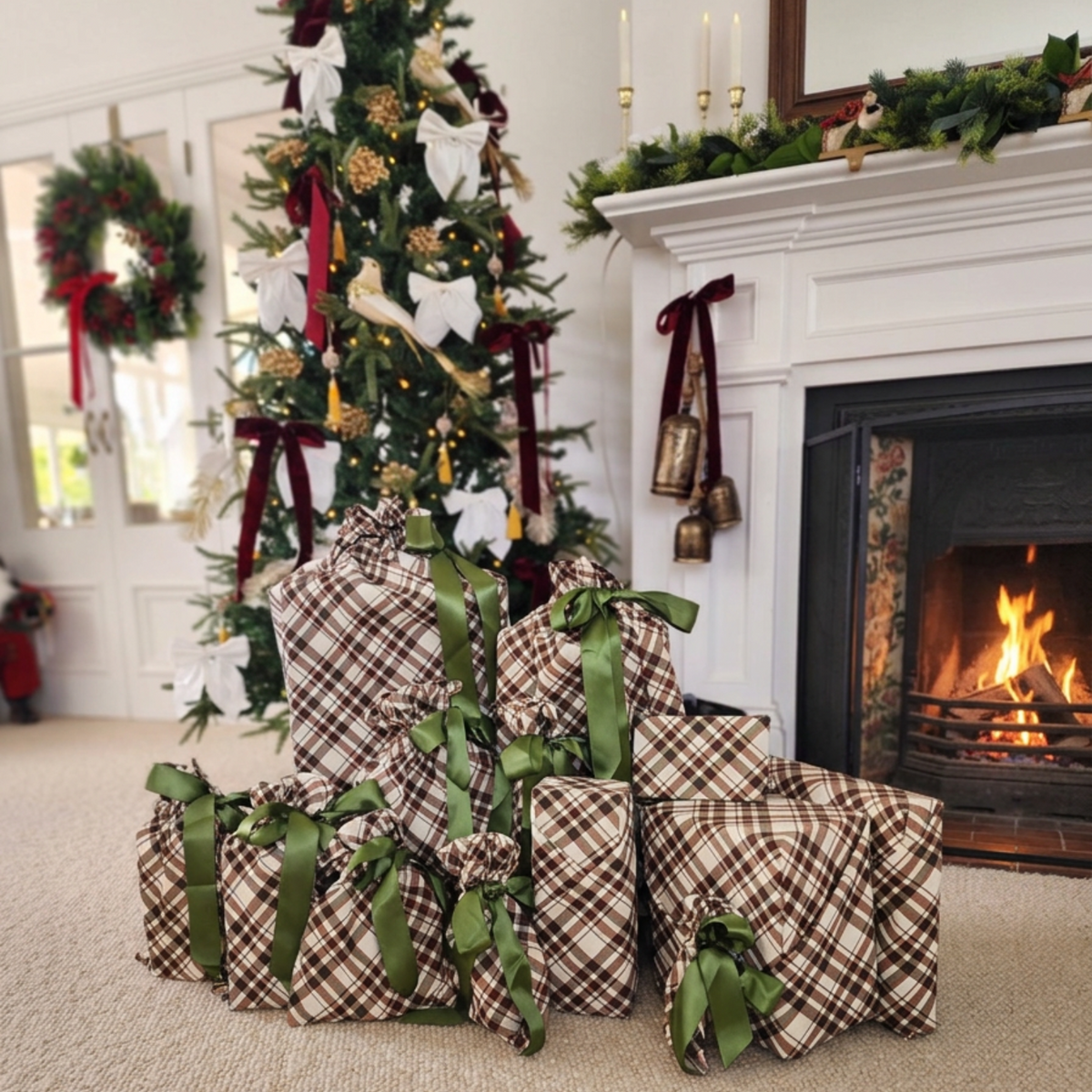 A group of wrapped Christmas presents in brown and beige plaid cotton with green ribbons, gathered on a carpeted floor in front of a decorated Christmas tree and a lit fireplace.