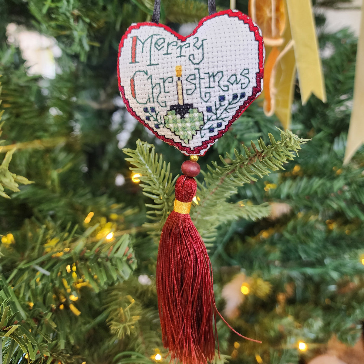 A heart-shaped embroidered Christmas ornament hanging on a green Christmas tree. The ornament has a white cross-stitch background with a red stitched border, the words 'Merry Christmas' in blue, a small yellow candle, and a green gift box.
