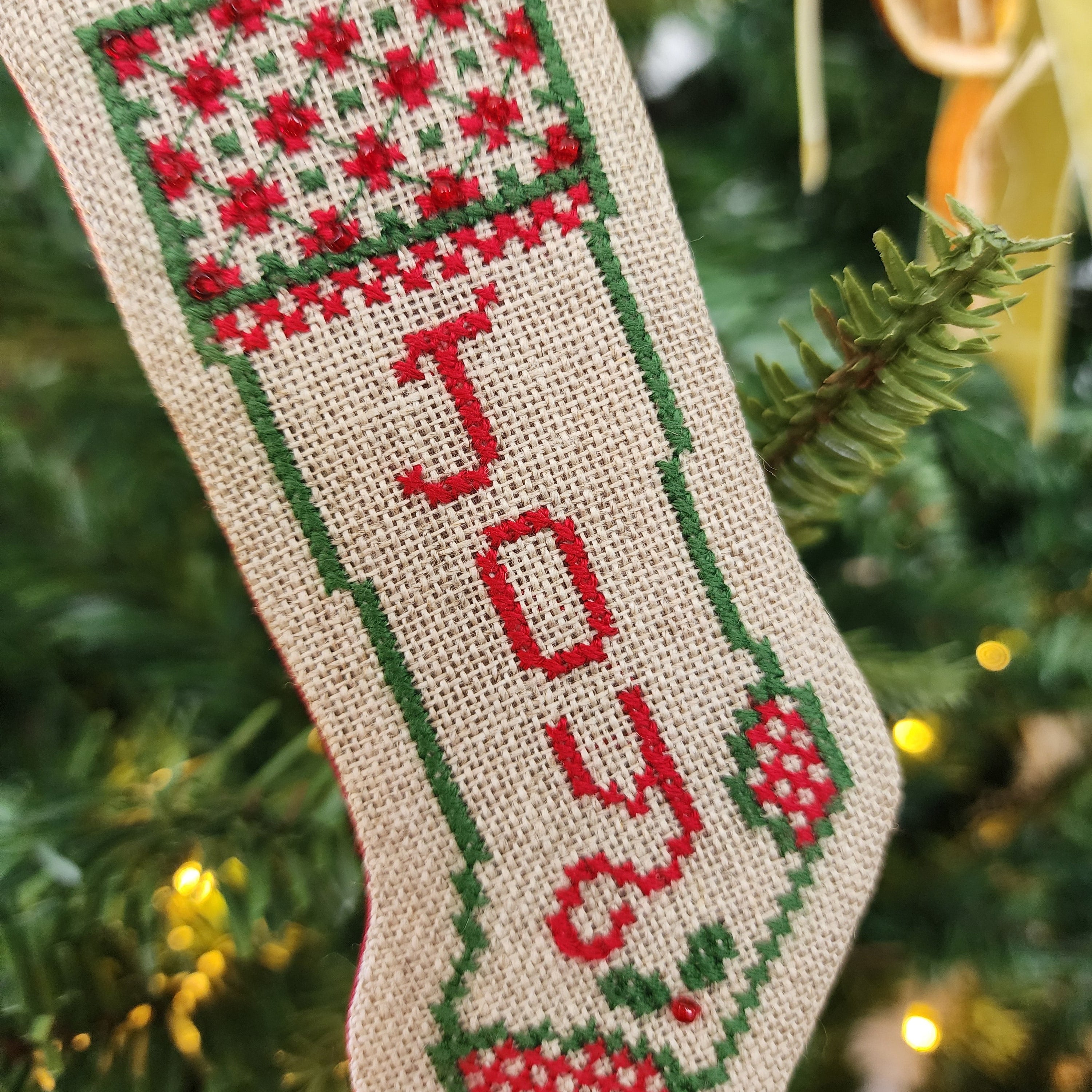 A handmade cross-stitch Christmas stocking ornament hanging on a decorated Christmas tree. The stocking is made of beige linen fabric and features red and green embroidery with the word 'JOY' stitched vertically in red.