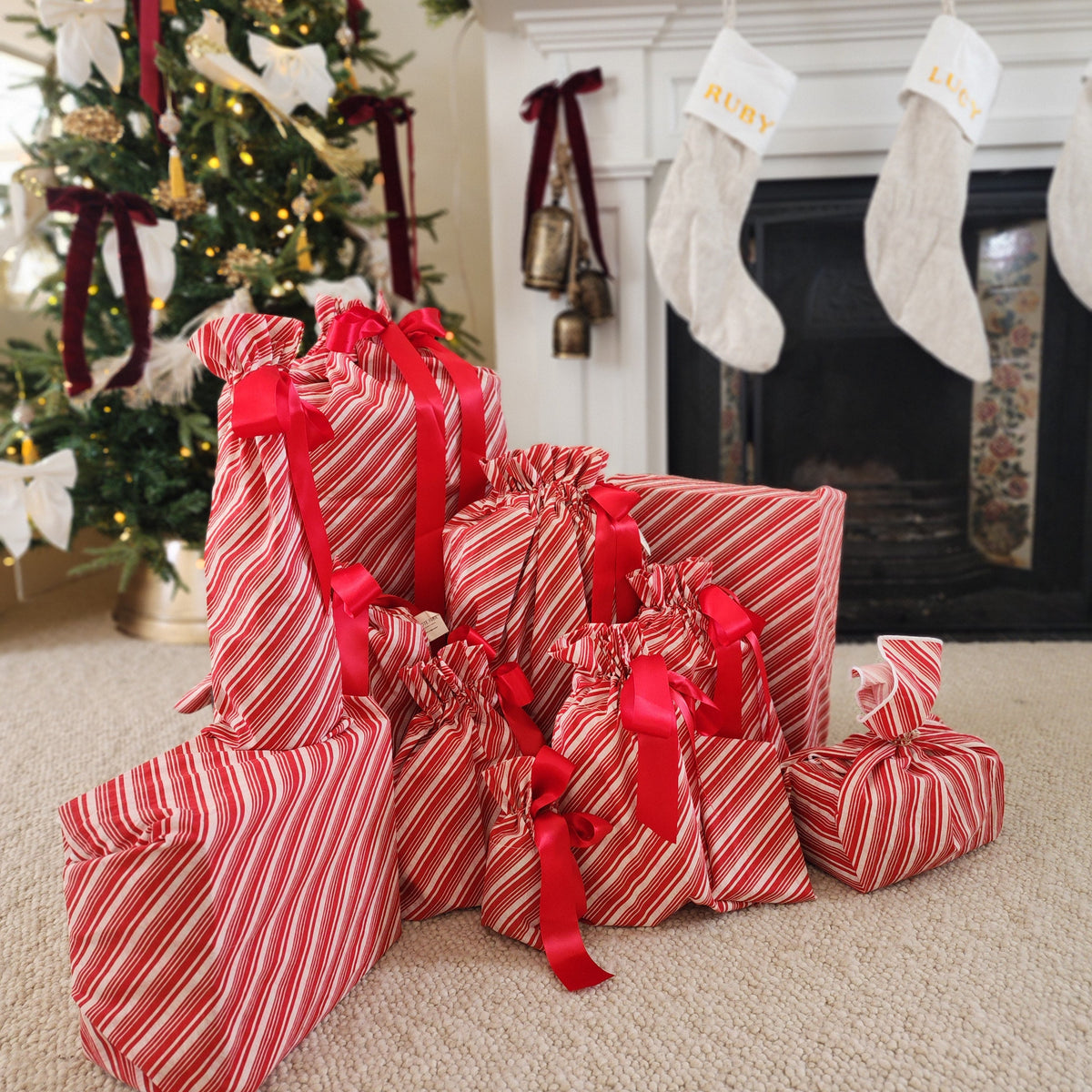 A group of Christmas wrapped gift boxes and stockings on a carpeted floor in front of a decorated fireplace and Christmas tree.