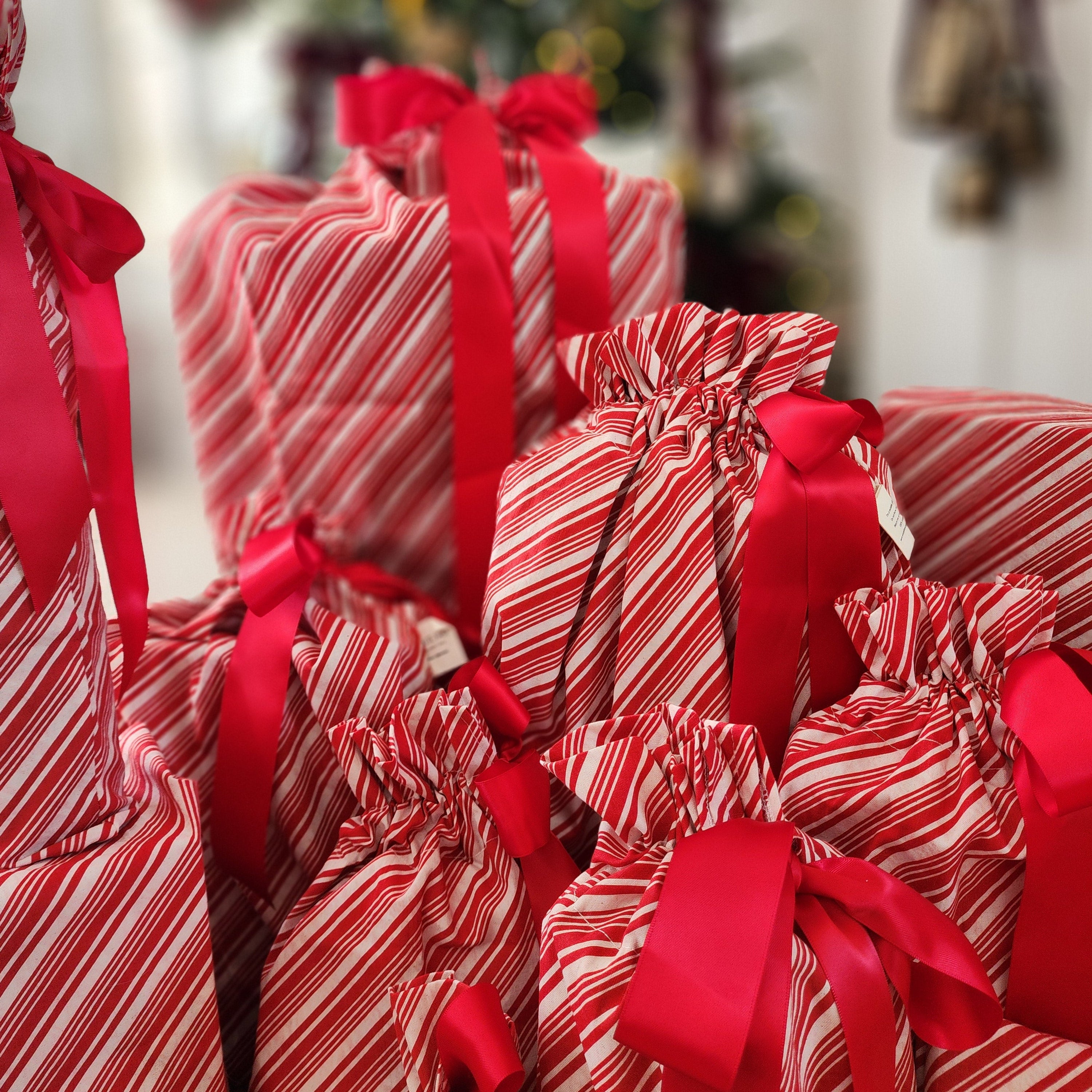 A close-up of multiple Christmas gift bags wrapped in red and white striped candy cane paper, tied with bright red satin ribbons, with a blurred decorated Christmas tree in the background.