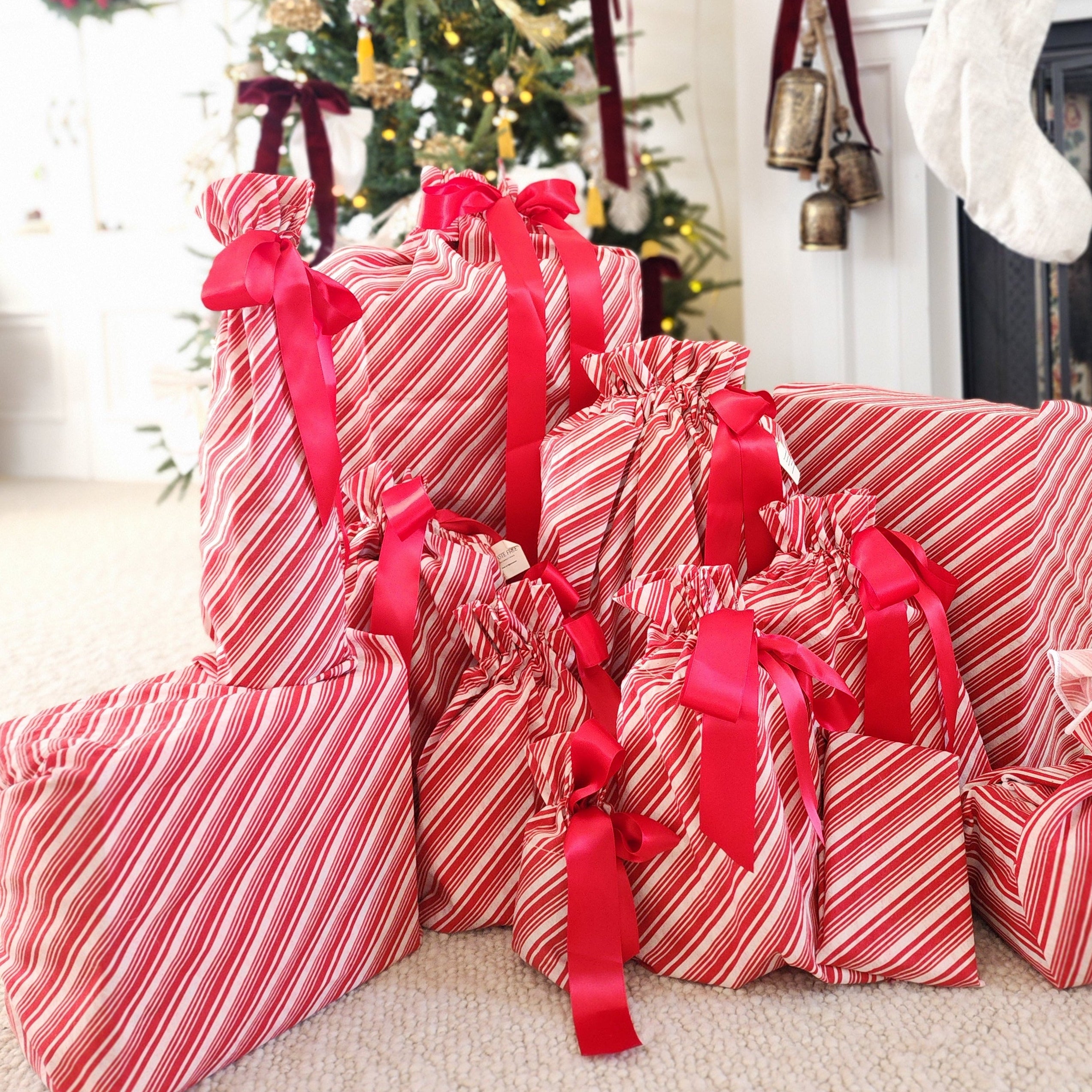 A collection of Christmas wrapped gift boxes and packages in red and white striped paper with red ribbons, arranged on a carpeted floor in front of a decorated Christmas tree and fireplace mantel.