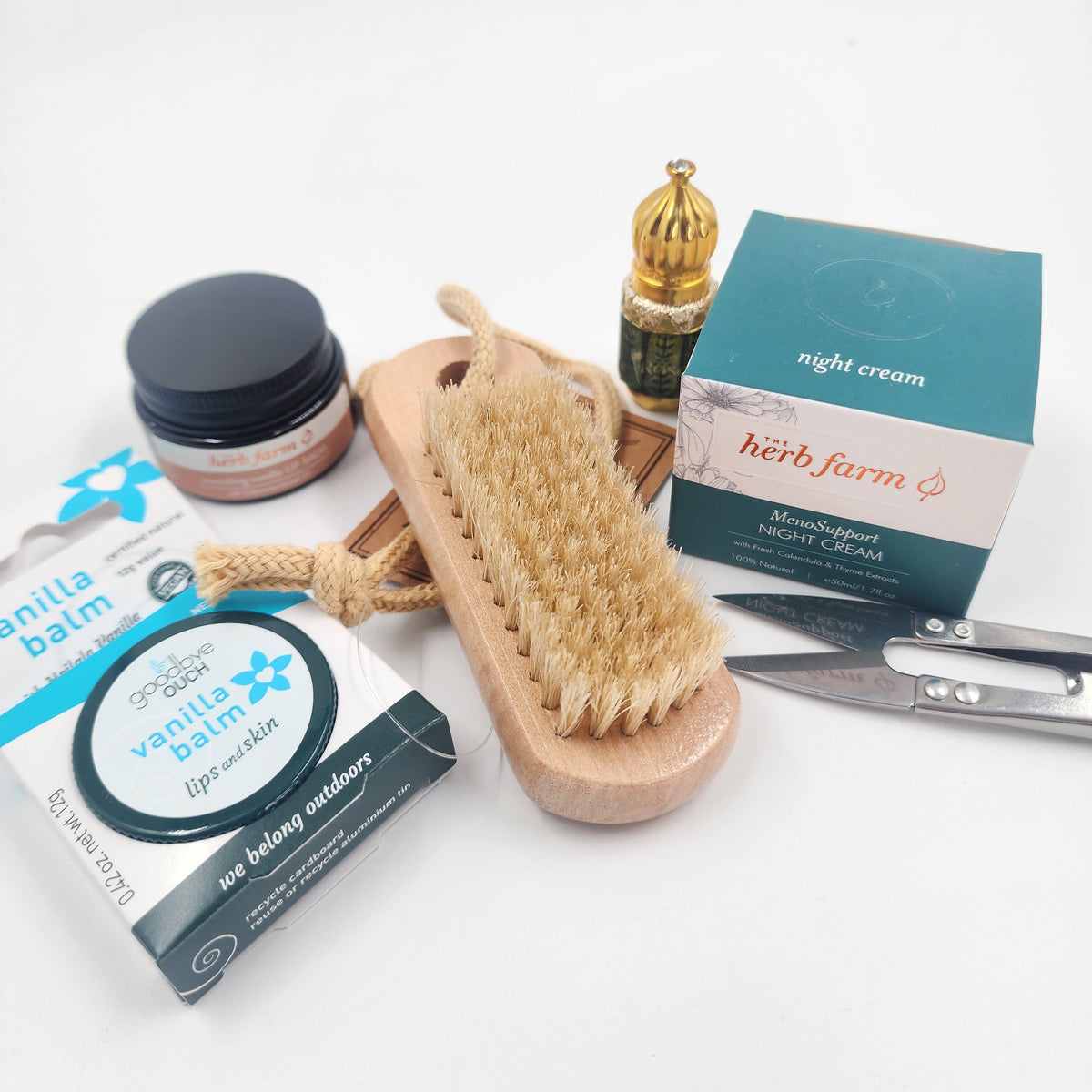 A curated beauty gift set on a white background, featuring a wooden hair brush with natural bristles, a jar of lip balm, a box of 'vanilla balm', a small gold-capped bottle, a teal and white 'herb farm' night cream box, and silver scissors.