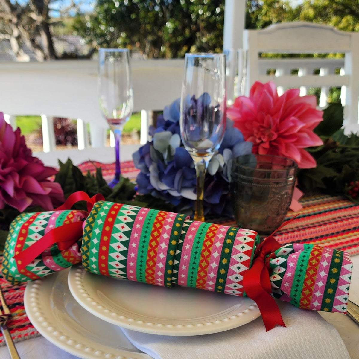 A festive outdoor table setting featuring a wrapped Christmas cracker with colorful geometric patterns and red ribbons, placed on a white tablecloth with plates, wine glasses, and flowers.