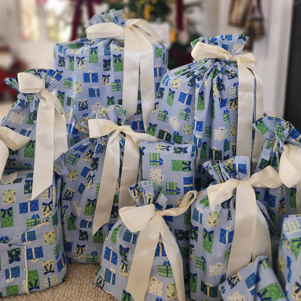 A group of Christmas or Hanukkah gift boxes wrapped in light blue fabric wrapping with a green and white holiday print, tied with cream-colored satin ribbons, set against a blurred festive background with a Christmas tree and fireplace.