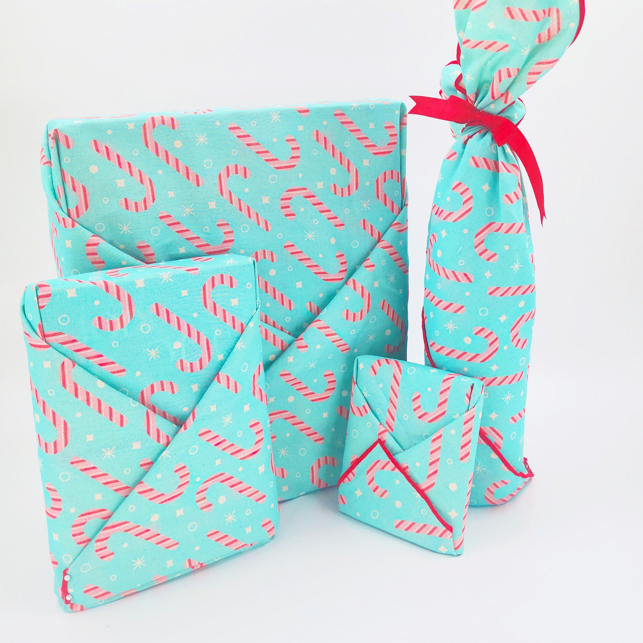 Three wrapped rectangular packages in matching turquoise furoshiki with a pink and white candy cane pattern, arranged on a white background. One package is tied with a bright red ribbon.