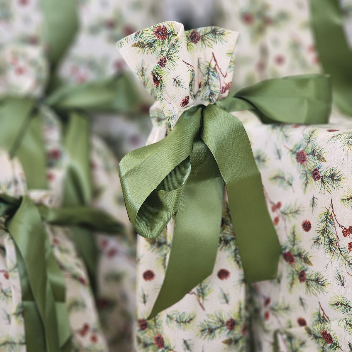 A close-up of a gift-wrapped package featuring a cream background with a green pine and berry pattern, tied with a wide olive green satin ribbon bow.