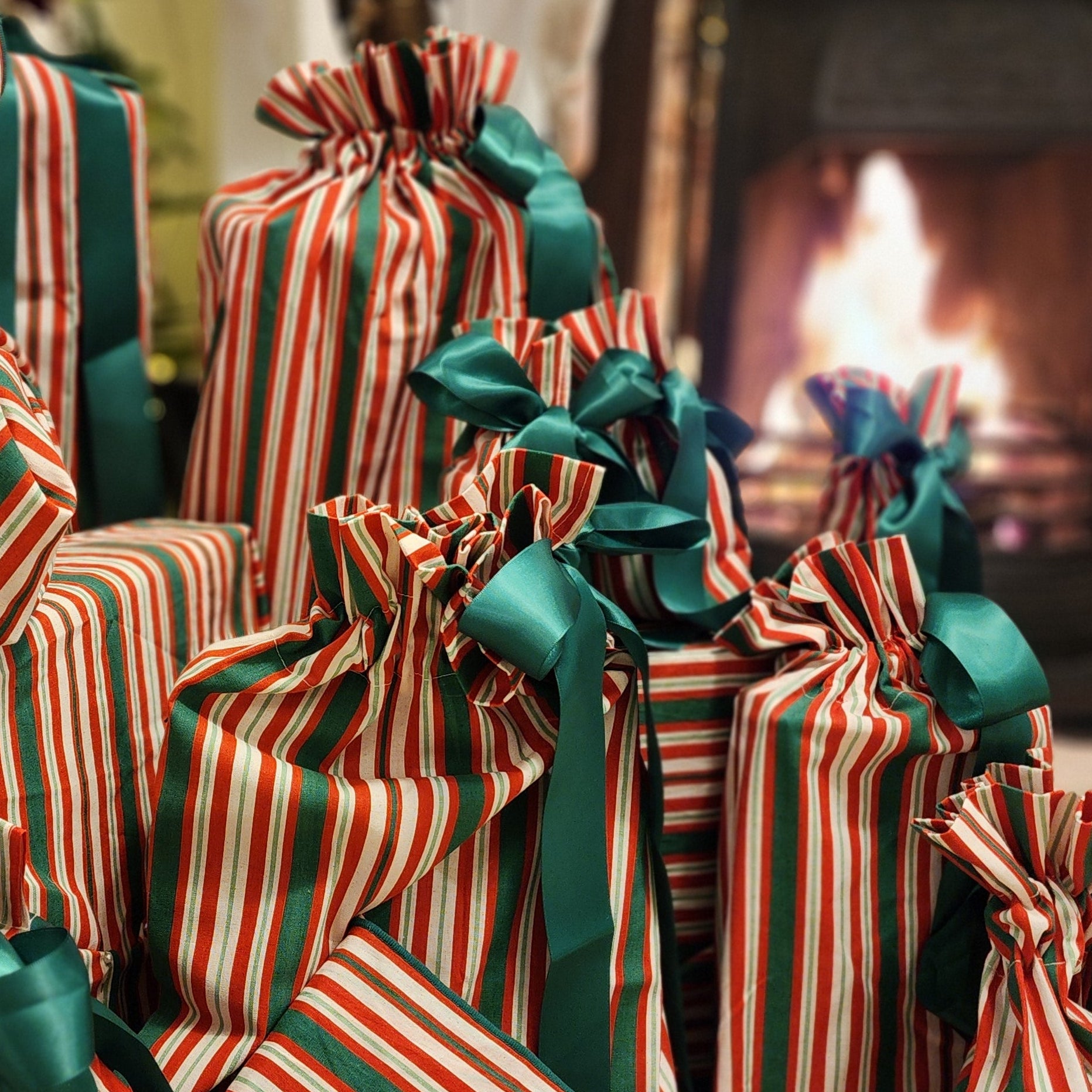 A group of Christmas gift bags wrapped in red, white, and green striped ribbon and tied with dark green satin bows, arranged on a carpeted floor in front of a fireplace with a lit fire and a decorated Christmas tree in the background.