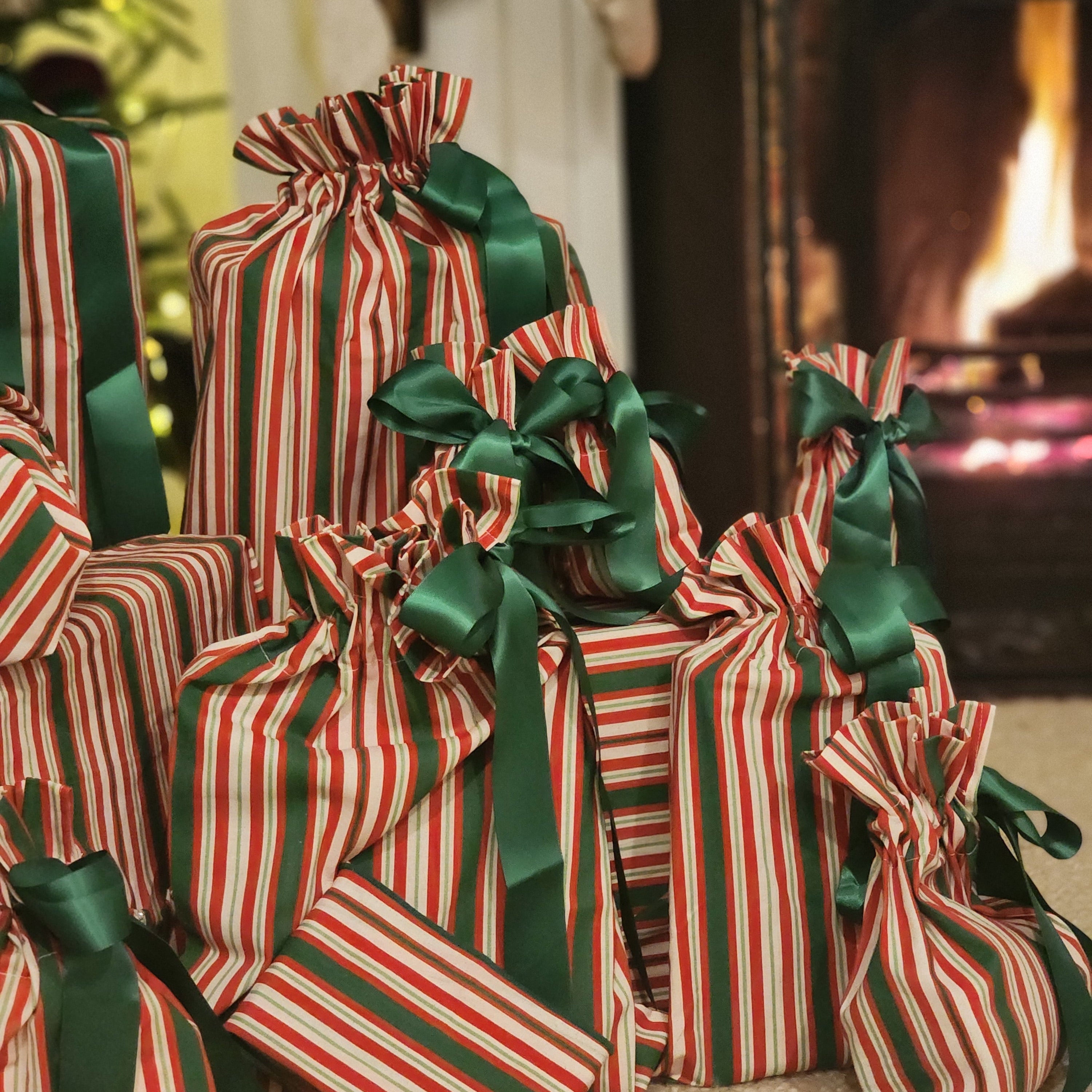 A group of Christmas-themed gift bags wrapped in red and white striped cotton bags with dark green satin ribbons and bows, arranged on a carpeted floor in front of a fireplace.