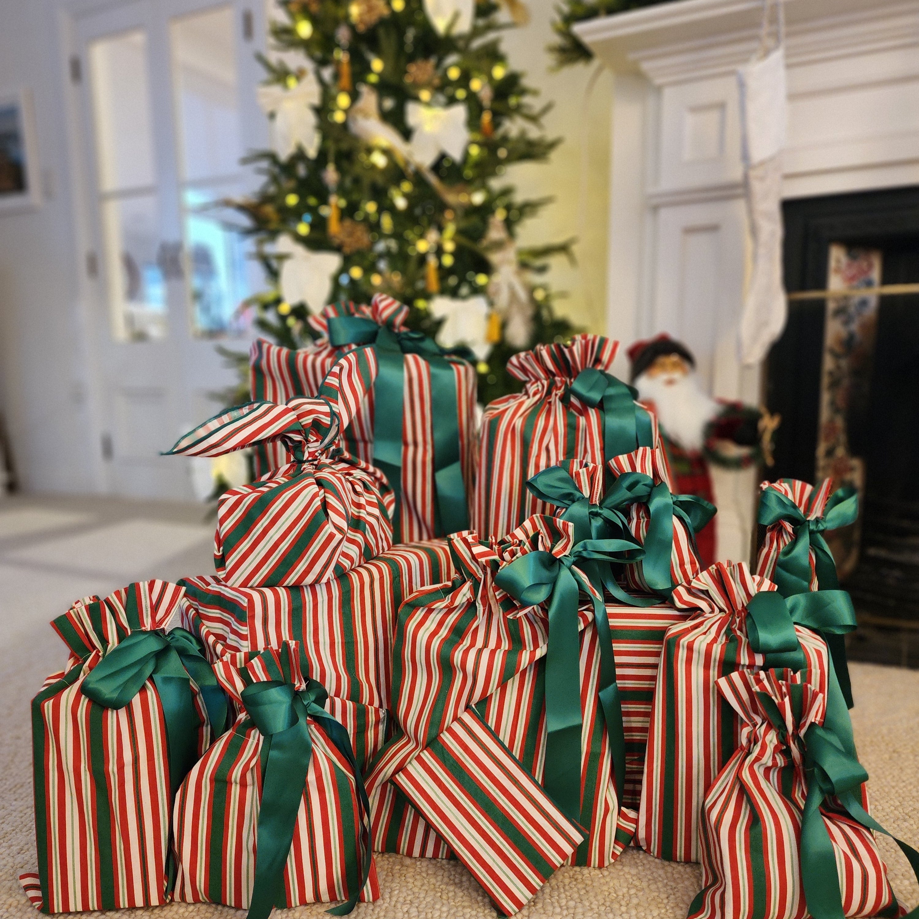 A group of small Christmas gift bags wrapped in red and white striped cotton bags and tied with green ribbons, arranged on a carpeted floor in front of a decorated Christmas tree and fireplace.