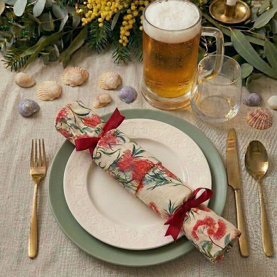 An Australian summer styled Christmas  table setting featuring reusable christmas cracker a red ribbon, layered white and sage green plates, gold flatware, a glass of beer, and seashells on a linen tablecloth.
