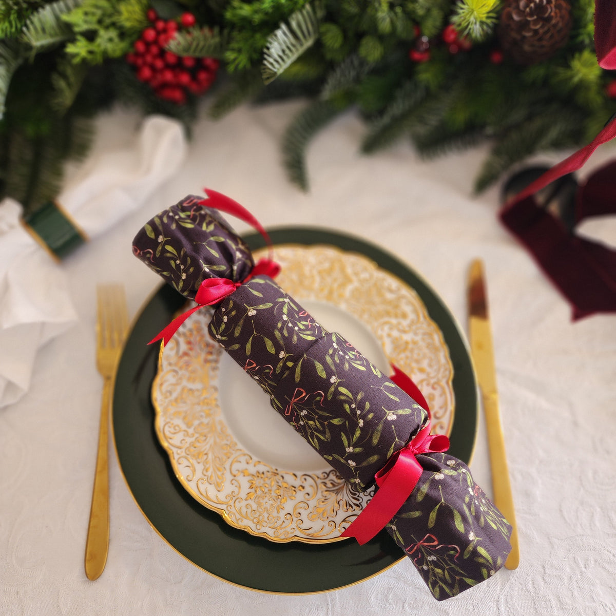 A festive Christmas table setting featuring a dark green charger plate with a gold rim, a white plate with gold detailing, gold-toned cutlery, and a wrapped Christmas cracker with red ribbons, set against a green pine garland background.