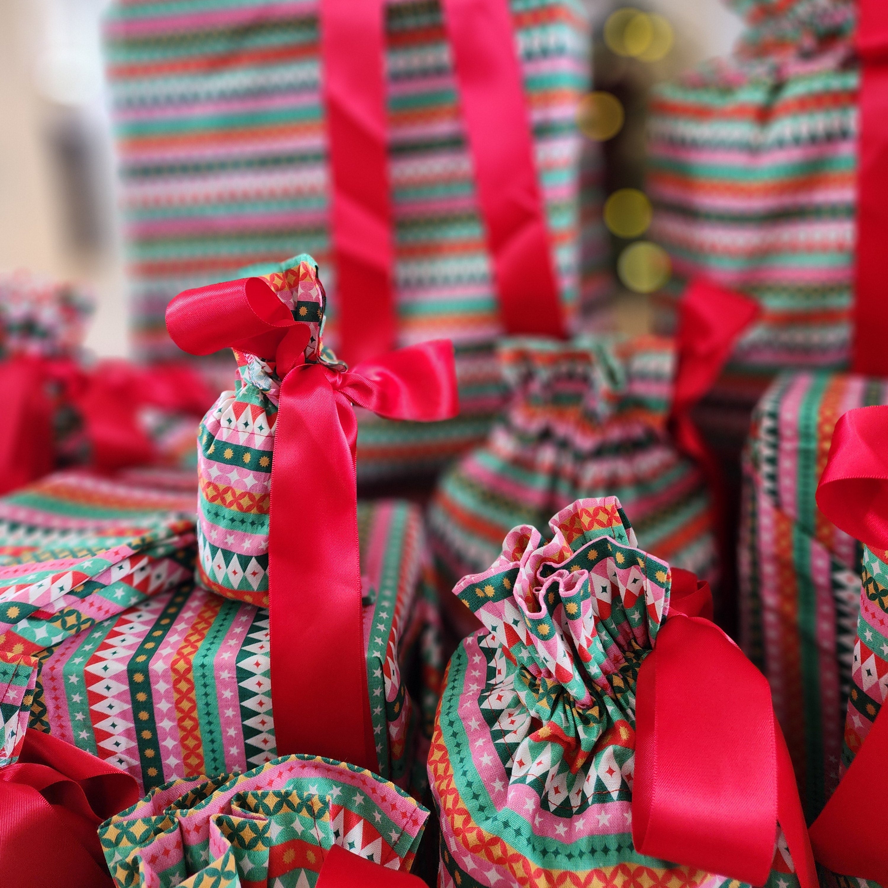 A collection of Christmas wrapped gift bags and boxes decorated with colorful geometric patterned cotton bags and bright red satin ribbons, set against a softly blurred festive background.