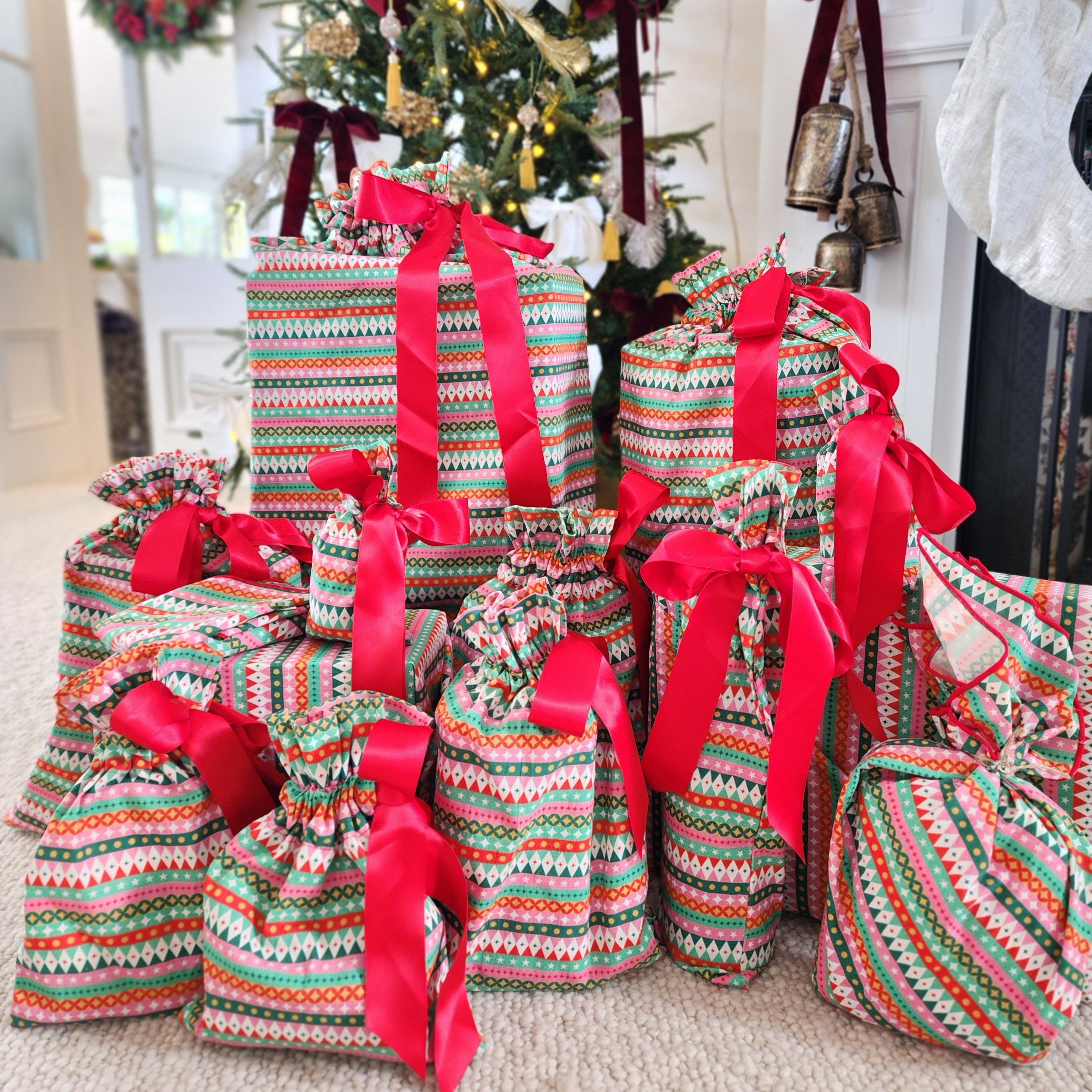 A collection of Christmas gift bags and wrapped presents filled with treats, decorated with red ribbons and multicolor geometric patterned paper, set on a carpeted floor in front of a decorated Christmas tree and fireplace mantel.