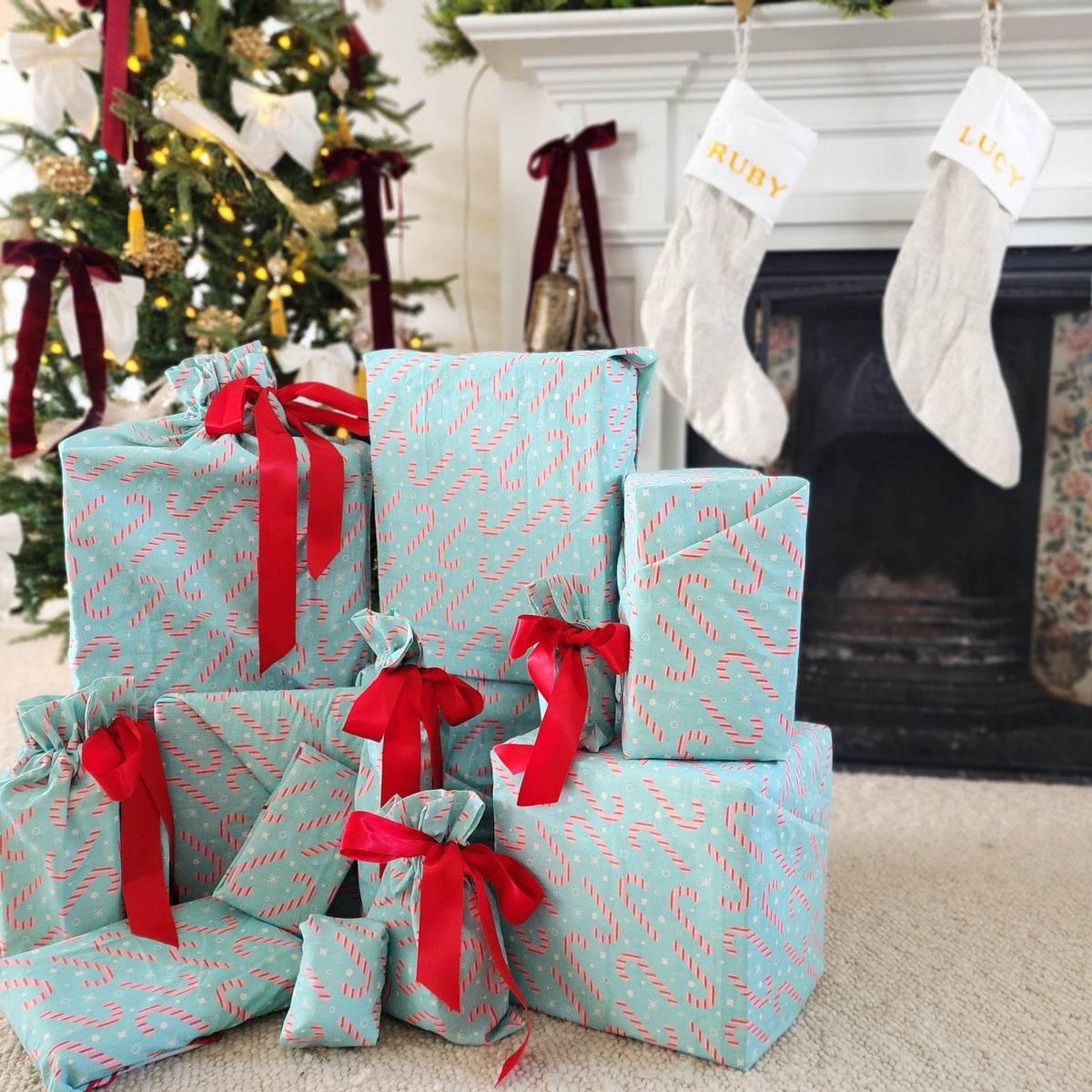 A collection of Christmas wrapped gifts in light blue candy cane patterned cotton gift bags and furoshiki with red ribbons, arranged on a beige carpet in front of a decorated fireplace with stockings.
