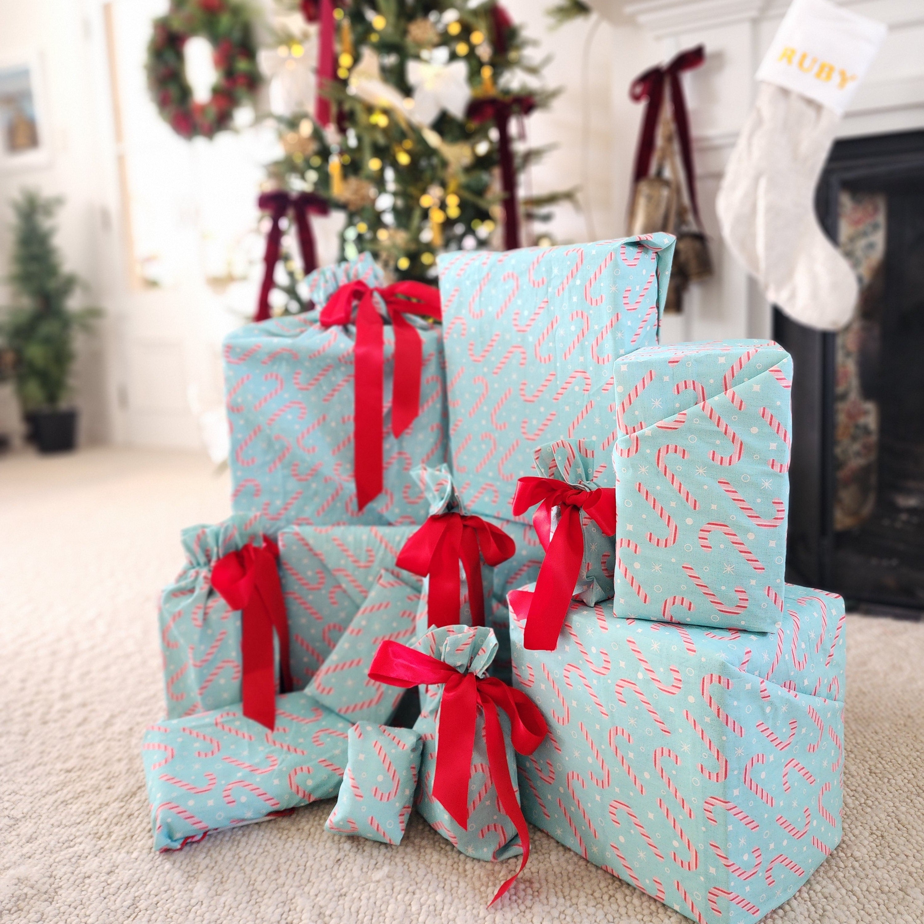 A collection of Christmas wrapped gifts in light blue cotton gift bags and furoshiki with a red candy cane pattern, tied with bright red ribbons, arranged on a beige carpet in a decorated living room with a Christmas tree and fireplace in the background.
