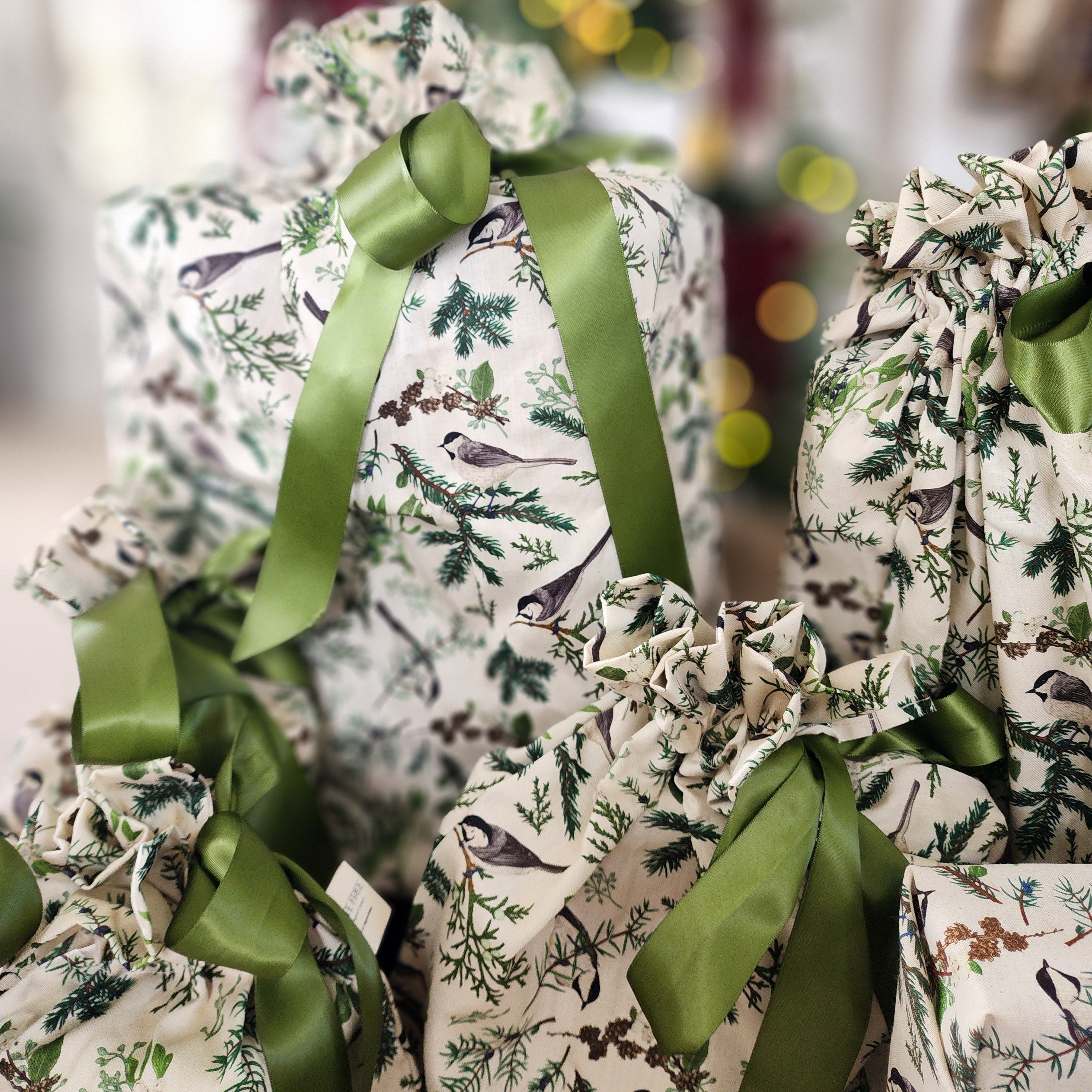 Christmas-themed gift bags and wrapped presents featuring a bird and pine branch print on a cream background, tied with olive green satin ribbons, with a blurred festive background.
