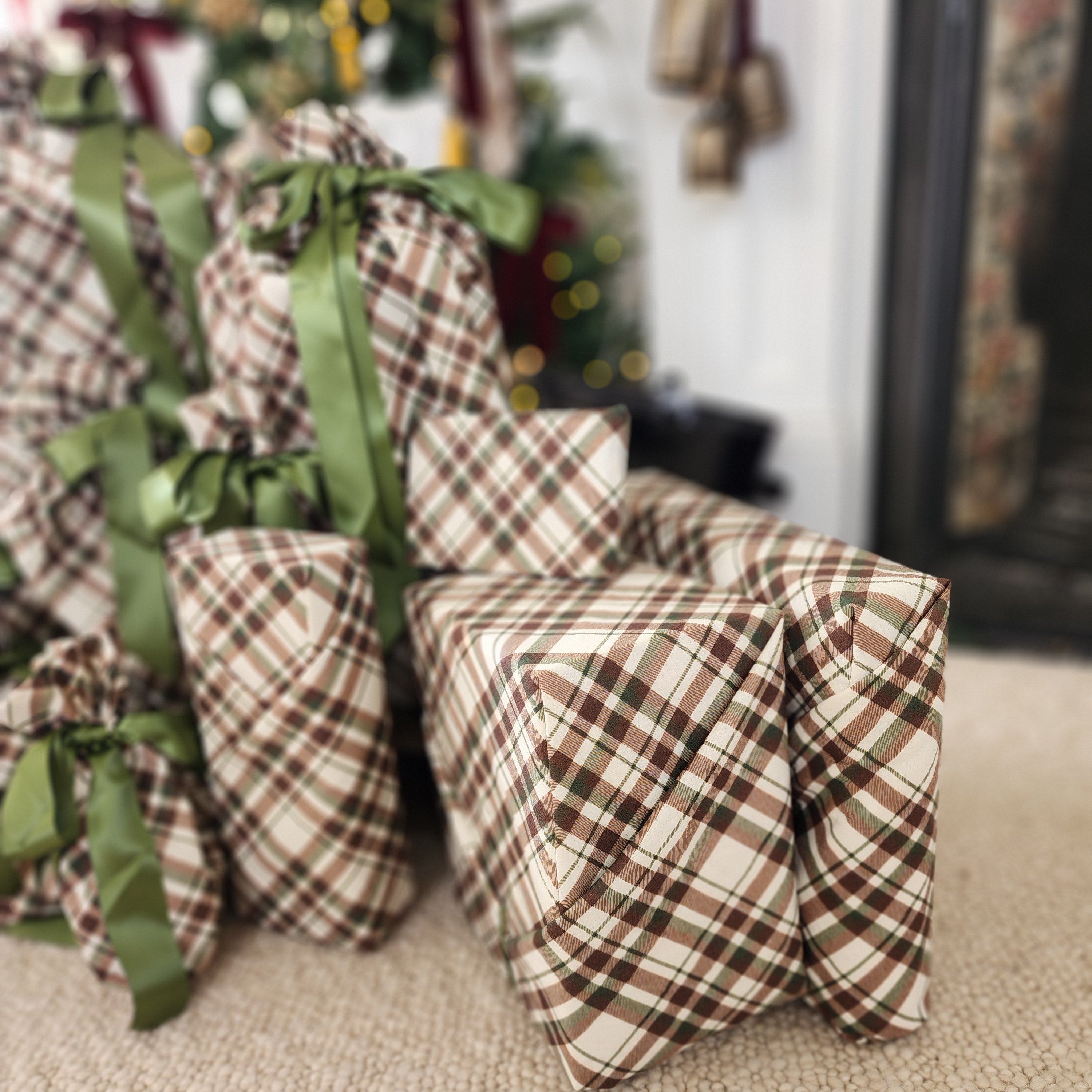A stack of Christmas wrapped gift boxes wrapped in brown, beige, and white plaid cotton with green ribbons, set on a beige carpet in a living room with a blurred Christmas tree in the background.