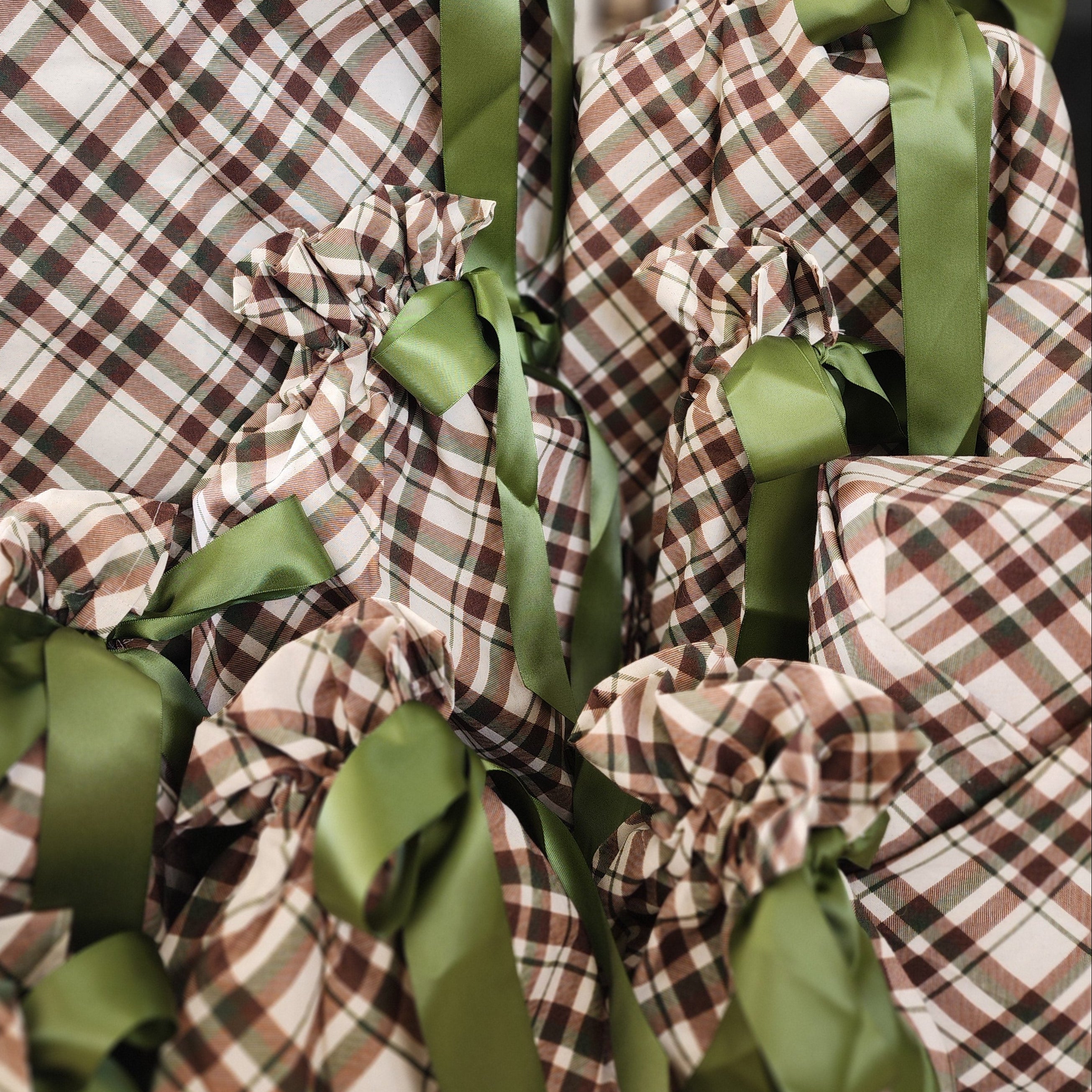 A group of Christmas wrapped gifts in brown, beige, and black plaid cotton with sage green satin ribbons tied in bows, set against a softly blurred holiday background.