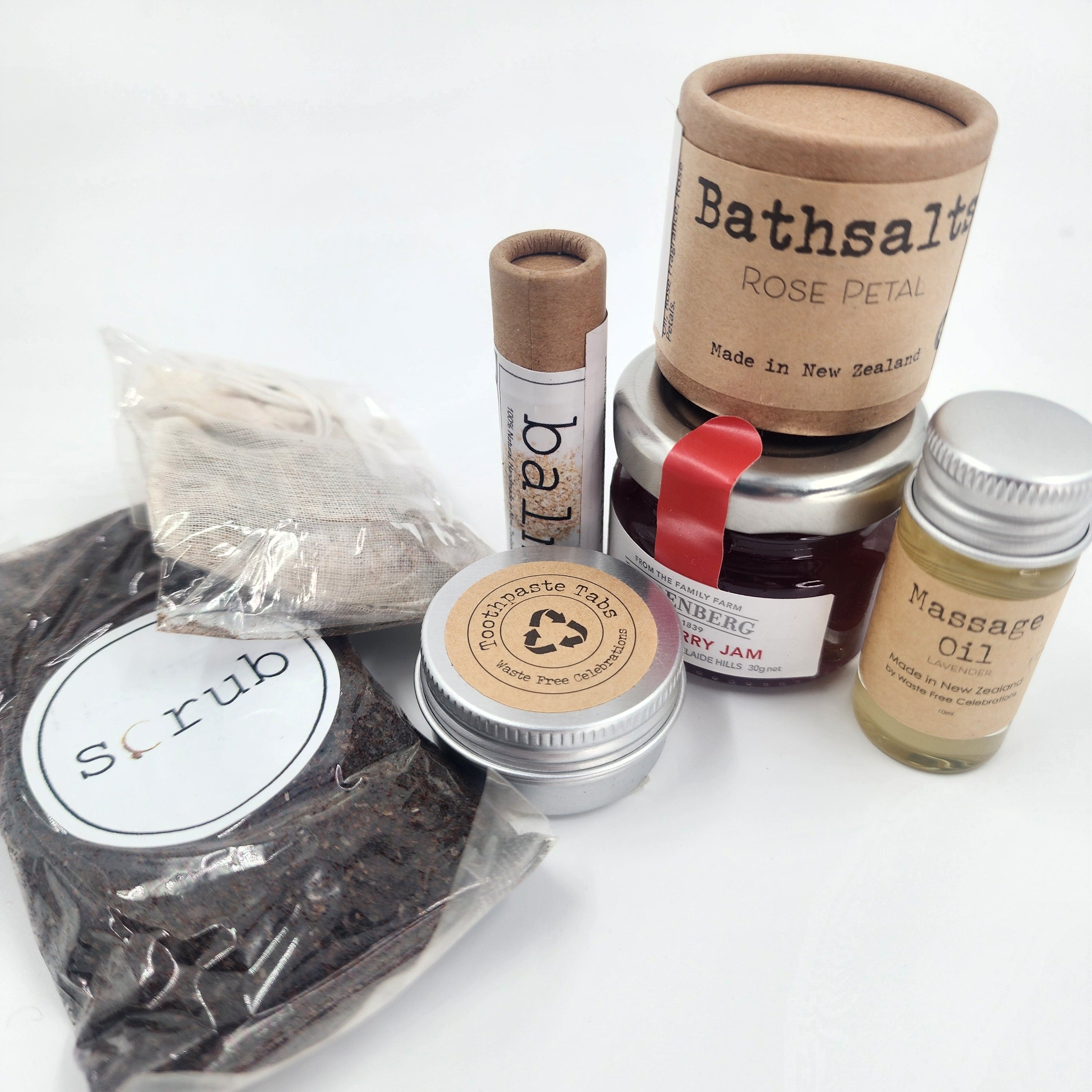 A bath and body gift set featuring a bag of dark brown herbal scrub, a tube of lip balm, a tin of jam, a jar of massage oil, and a large cardboard canister of rose petal bath salts, arranged on a white background.