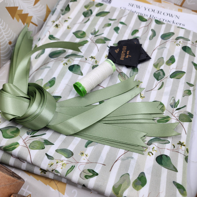 Closeup photo of Australiana Watercolour fabric, with green satin ribbon, tags and thread. The fabric has taupe and white stripes with green leaves on it.