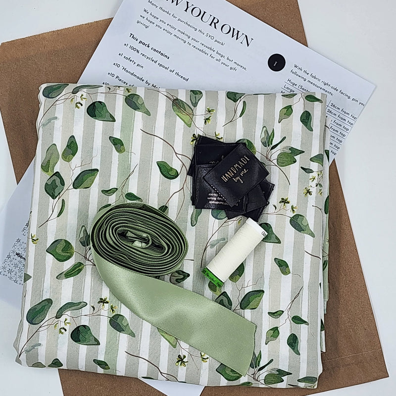 A photo showing the contents of a Sew Your Own gift Bag set in Australiana Watercolour. There is fabric, instructions, satin ribbon, tags and thread. The fabric has taupe and cream stripes with a green leaf print.