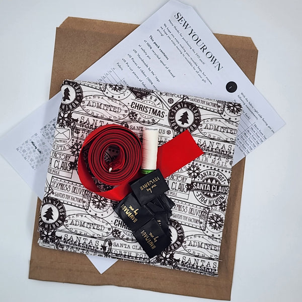 A photo showing the contents of the Sew Your Own Gift Bag set in Santas Mail. There is fabric, satin ribbon, thread, tags and instructions. The fabric is white with black postage stamps on it.