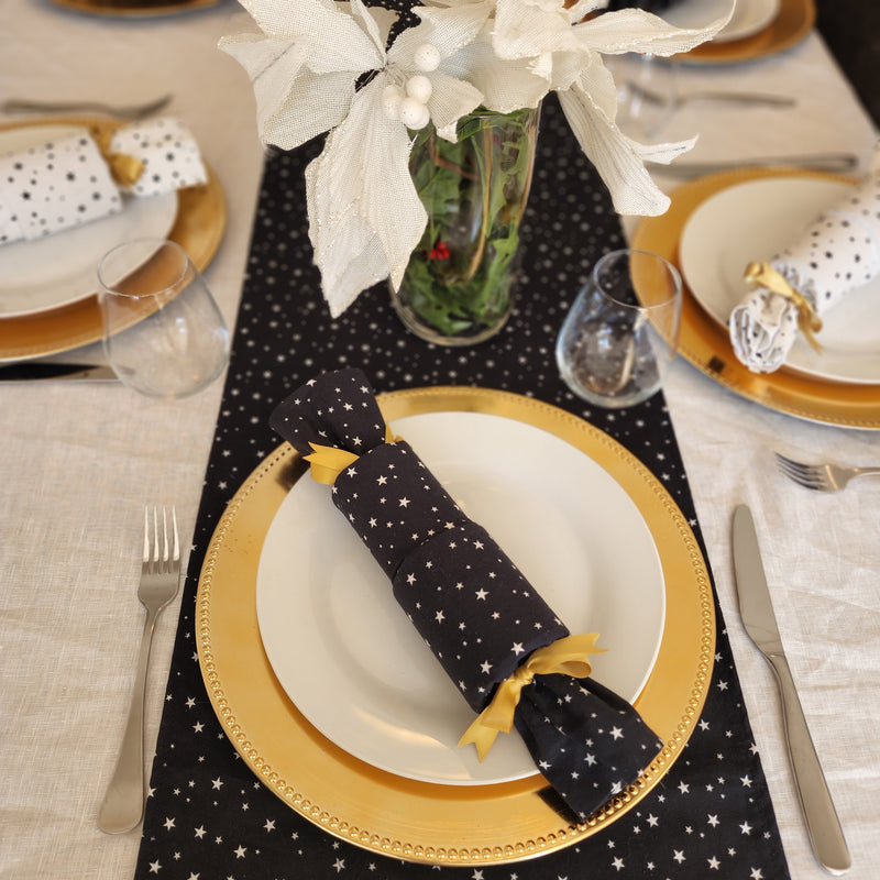 The image is of a table setting using Starry Night Re-Crackers (reusable Christmas Crackers or Bon bons). The crackers are white with tiny black stars, or black with tiny white stars,  and is finished with a gold ribbon. The crackers are sitting on plates and the table is set for Christmas.